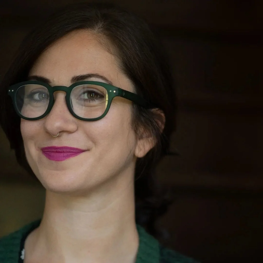 Close-up of a woman with short dark hair, wearing green glasses, pink lipstick, and a nose septum piercing, smiling against a dark background.
