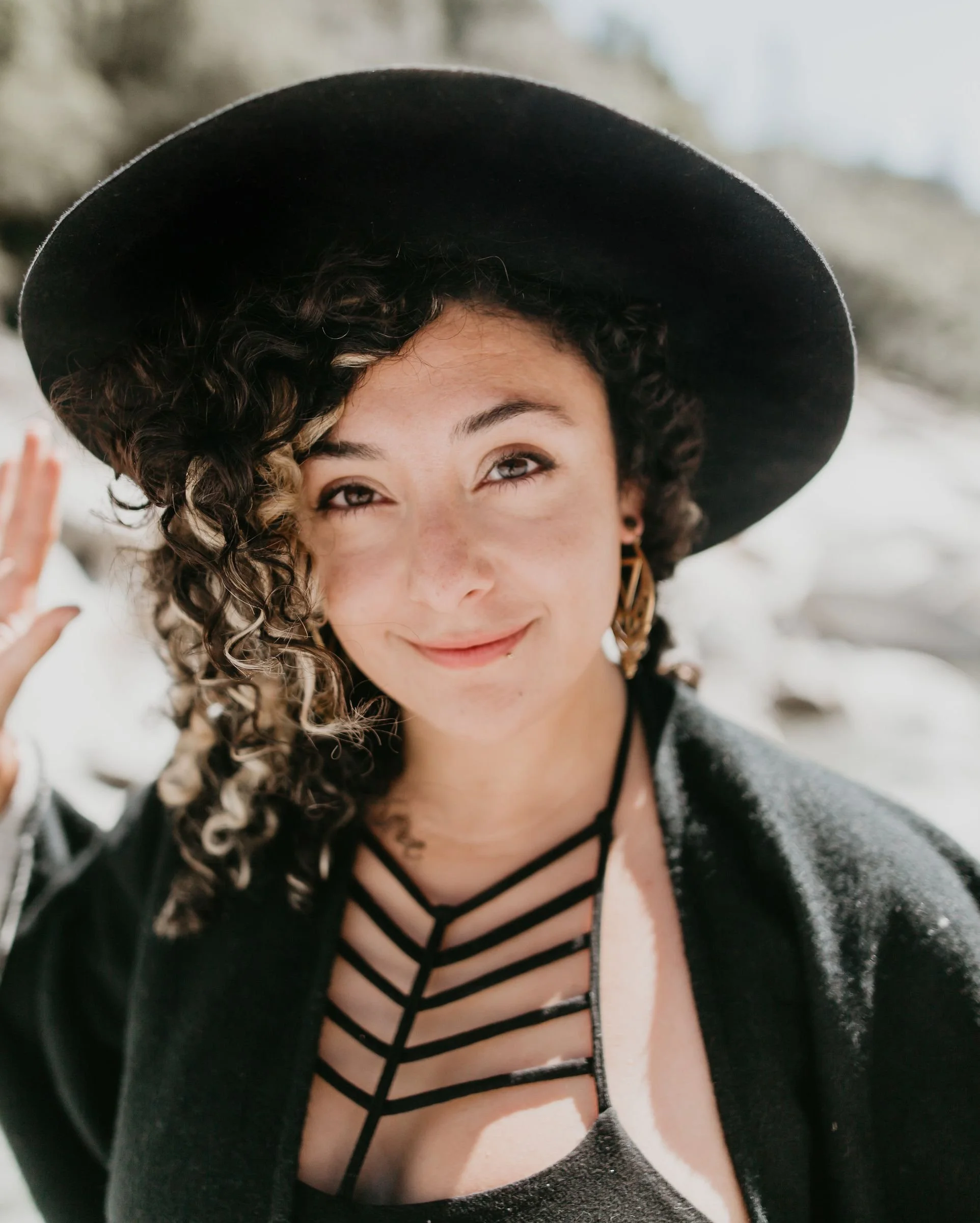 A woman with curly hair wearing a black hat and a black top with a crisscross design, outdoors with snow and rocks in the background, smiling and waving.