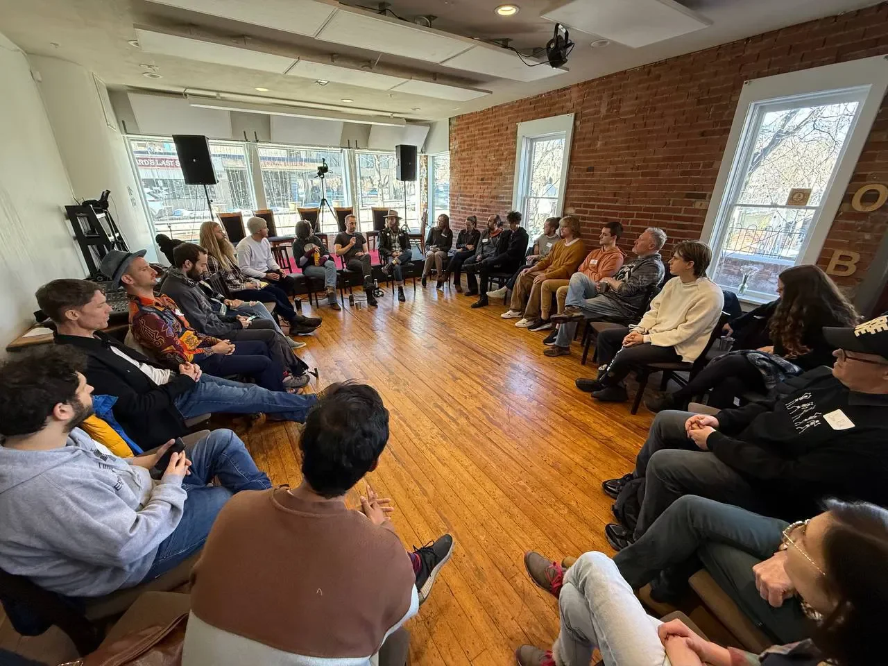 A group of people sitting in a circle in a bright room with wooden floors and brick walls, participating in a discussion or workshop.