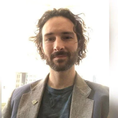 Portrait of a man with curly dark hair and a beard, wearing a blazer and a t-shirt, standing in front of a window with buildings outside.