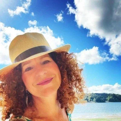 Woman with curly hair and a hat smiling outdoors near a lake with blue sky and clouds.