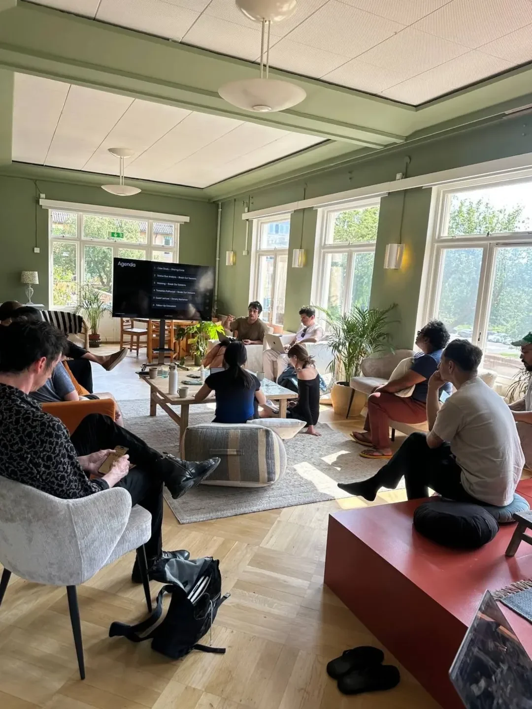 A group of people attending a presentation or meeting in a bright, spacious room with large windows, a television screen displaying an agenda, and various seating arrangements including armchairs, a low table, and cushions on the floor.