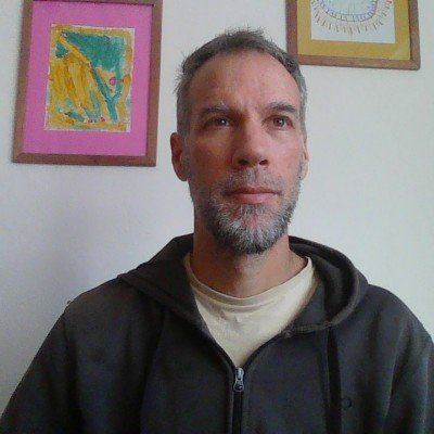 A man with short hair and a beard, wearing a black hoodie and a beige shirt, standing in front of a white wall with colorful children's artwork framed and hanging.