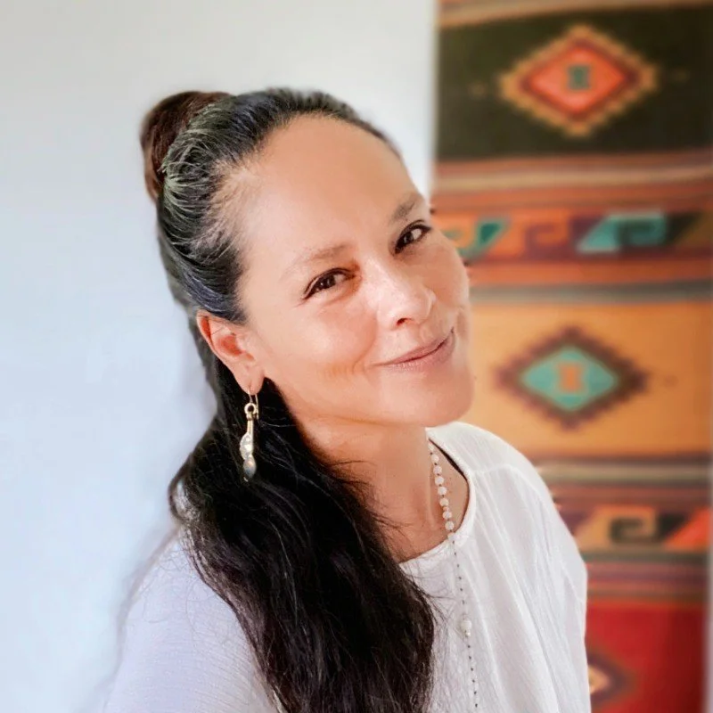 A woman with dark hair in a half-up hairstyle, wearing pearl earrings and a white top, smiling softly, standing against a colorful, patterned background.