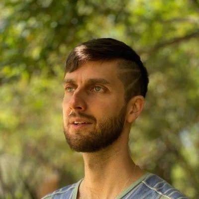 A young man with a beard and short brown hair standing outdoors in a forested area.