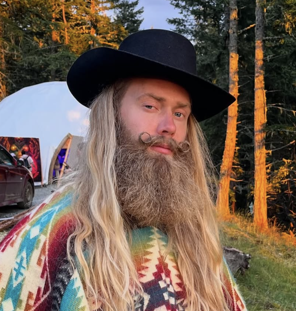 A man with long blonde hair and a large beard wearing a black wide-brimmed hat and a colorful, patterned poncho standing outdoors near trees at sunset.
