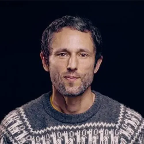 A man with short curly hair and a short beard, wearing a gray sweater with white snowflake patterns, against a dark background.