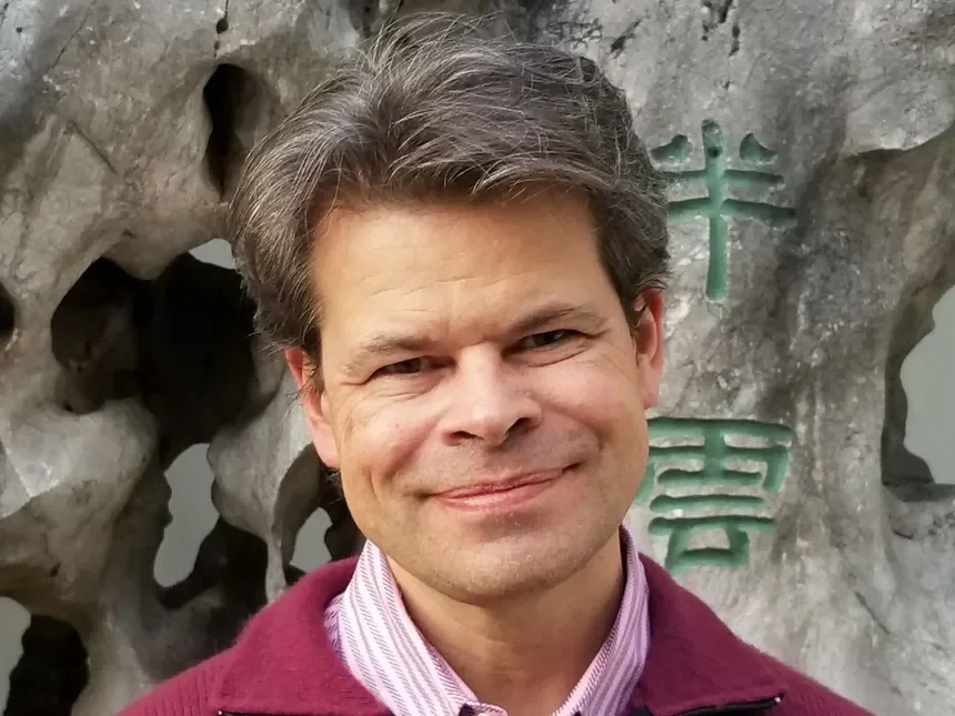 A smiling man with light brown hair standing in front of a large rock with green Chinese characters engraved on it.