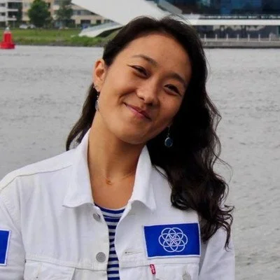 A young woman with long dark hair smiling by a body of water, wearing a white jacket with a blue logo and earrings.