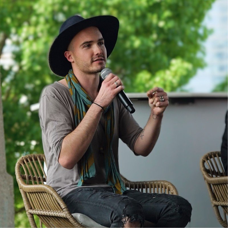 Man in a black wide-brim hat speaking into a microphone during an outdoor event with green trees in the background.
