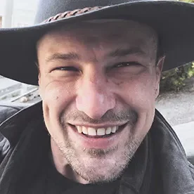 A smiling man wearing a wide-brimmed hat and a dark jacket outdoors.