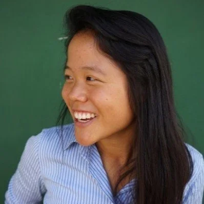 A woman smiling and looking to her left, with long dark hair and wearing a blue striped shirt, in front of a green background.