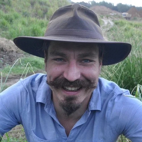 A man outdoors wearing a wide-brimmed hat and a blue shirt, smiling at the camera, with greenery and a dirt path in the background.