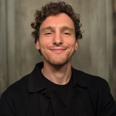 A man with curly hair and a beard smiling, wearing a black shirt against a neutral background.