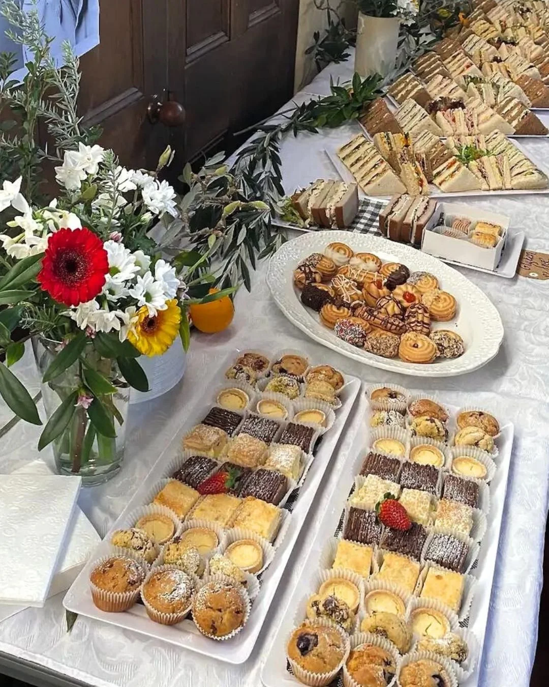 A table with an assortment of baked goods, sandwich trays, and a floral arrangement. The baked goods include muffins, cookies, and pastries, with some strawberries on top. Sandwiches are arranged on platters, and there's a vase of colorful flowers on the table.