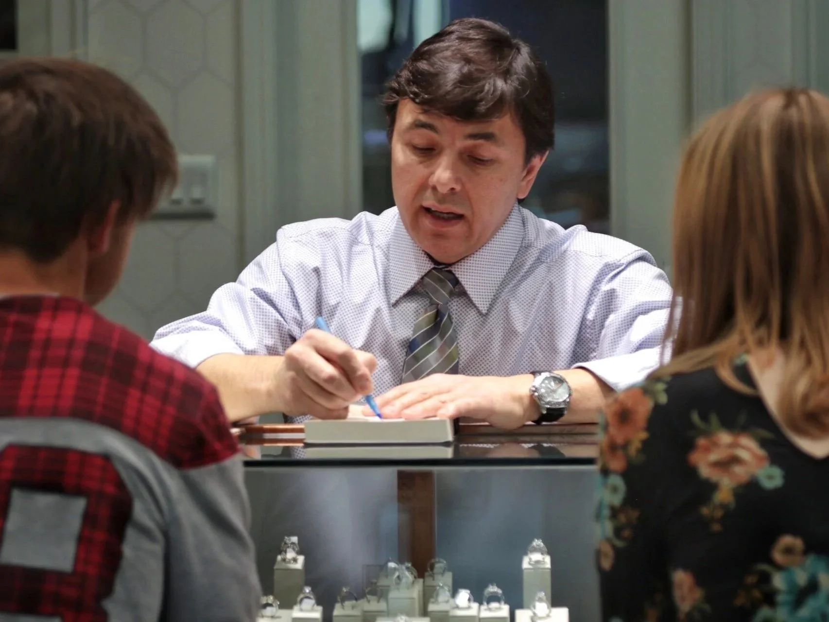 A man with dark hair wearing a white shirt and striped tie is sitting at a table, talking to a woman with light brown hair wearing a floral dress and a man with short brown hair and a red plaid shirt, while placing jewelry on display in a store.