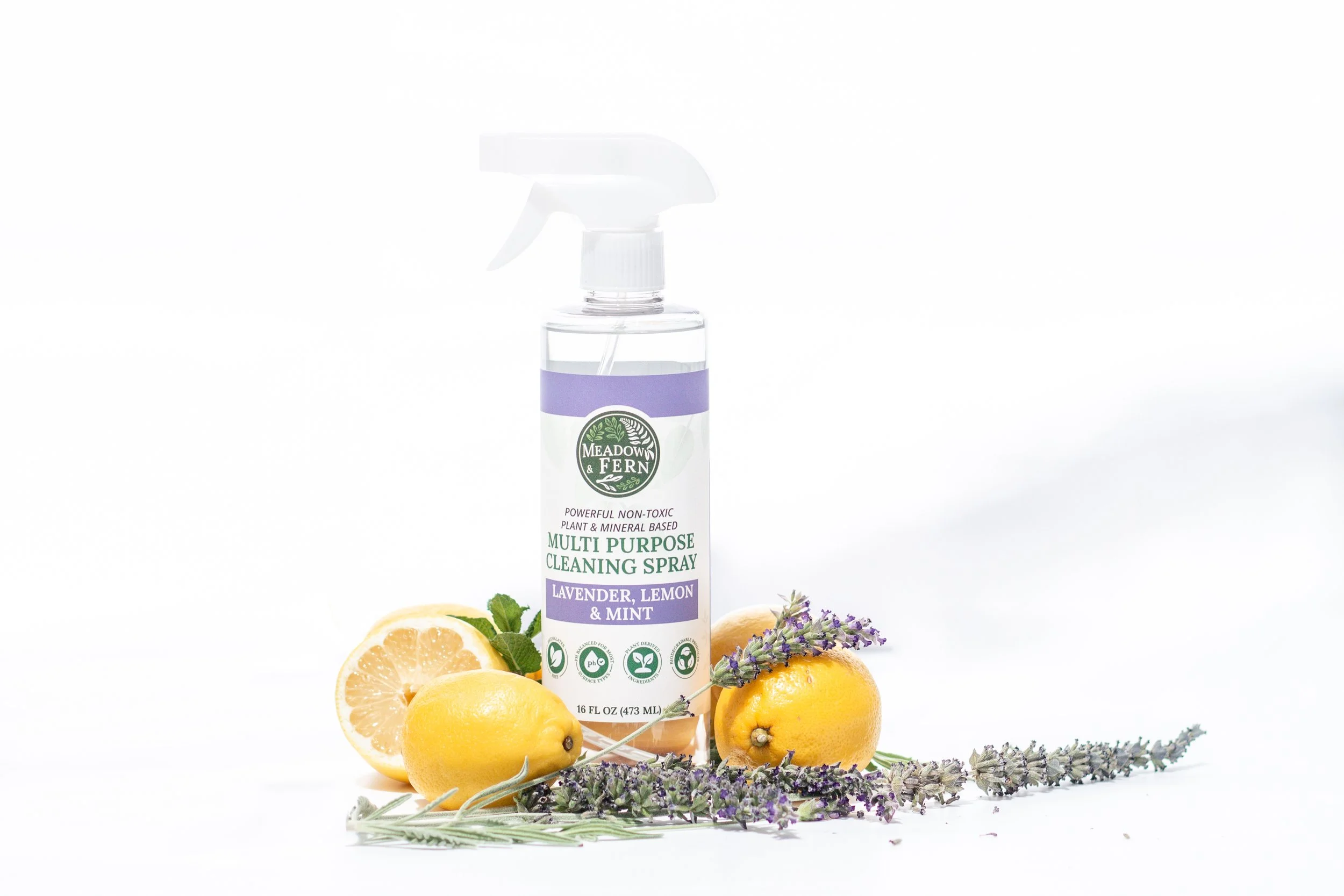 A bottle of Meadow & Fern multi purpose cleaning spray labeled 'Lavender, Lemon & Mint' surrounded by fresh lemons, lavender sprigs, and mint leaves on a white background.