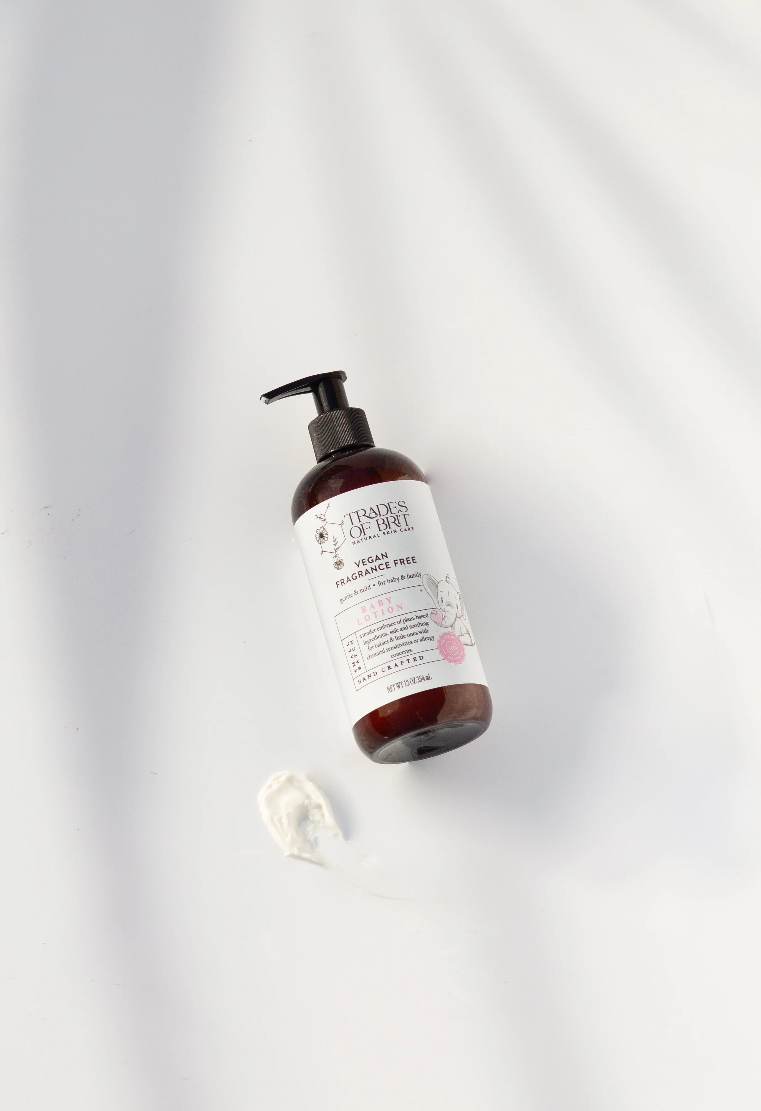 A bottle of Trades of Brit vegan fragrance-free baby lotion with a pump dispenser, lying on its side on a white surface, with a dollop of white lotion underneath.