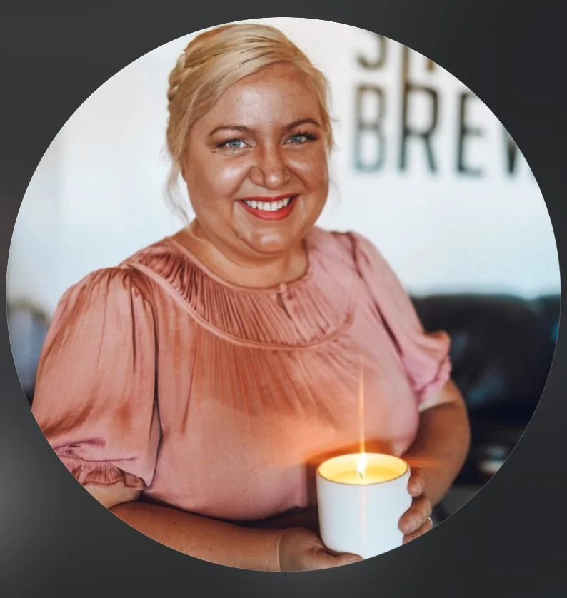 A woman with blonde hair smiling, holding a lit candle in a white mug, wearing a pink blouse with puffed sleeves, in an indoor setting.