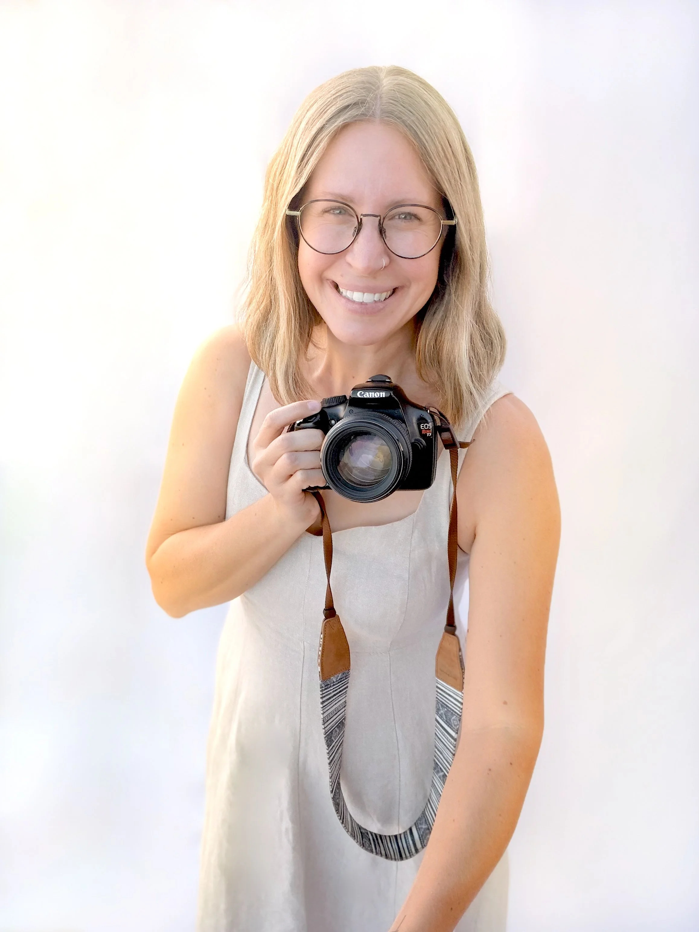 A woman with blonde hair, wearing glasses and a light-colored dress, smiling and holding a professional camera, standing against a plain light background.