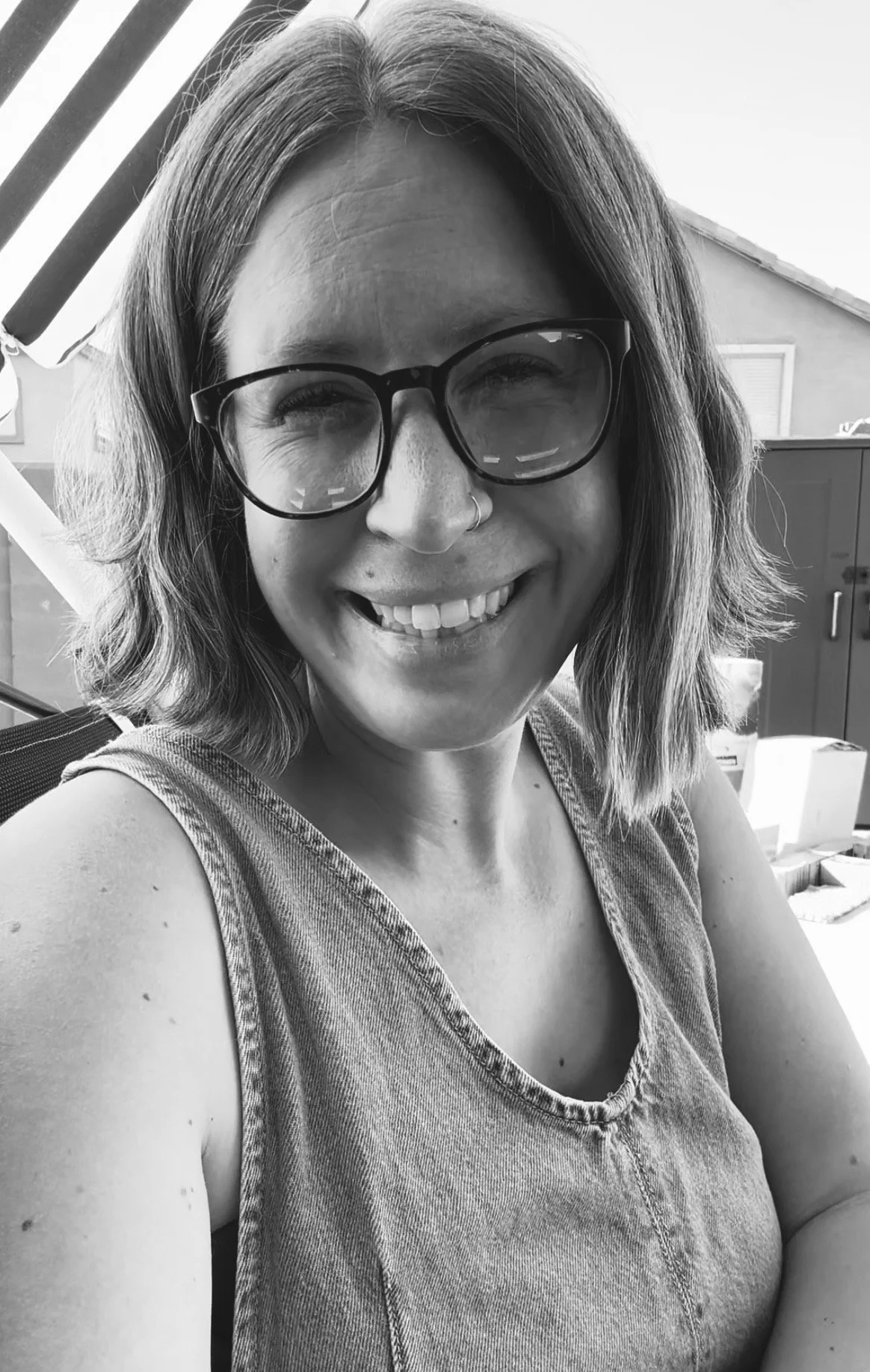 A woman with glasses and a nose ring smiling outdoors. She has shoulder-length hair and is wearing a sleeveless top.