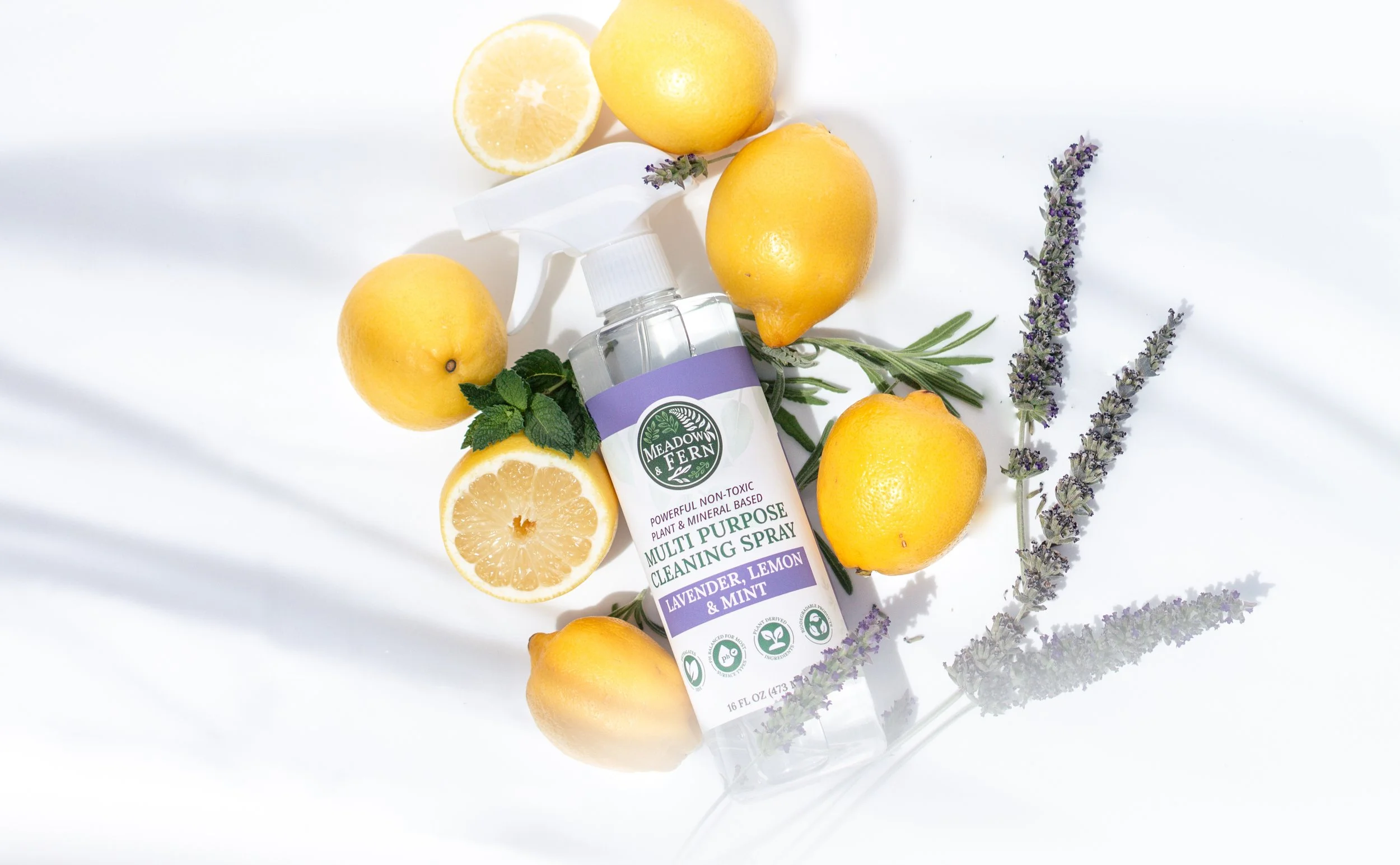 A Meadow Fern multi-purpose cleaning spray bottle labeled lavender, lemon, and mint surrounded by fresh lemons, lemon slices, sprigs of mint, and lavender on a white background.