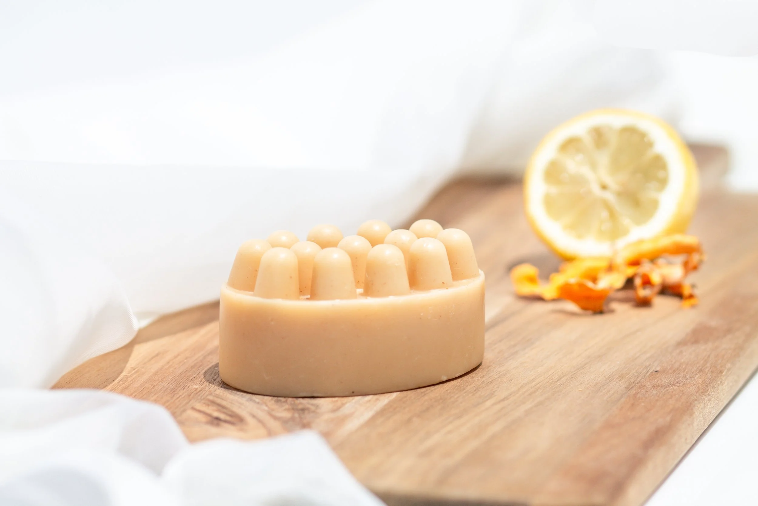 A bar of soap with a rounded top and multiple small, rounded protrusions on top, placed on a wooden surface next to a halved lemon and some orange peel.