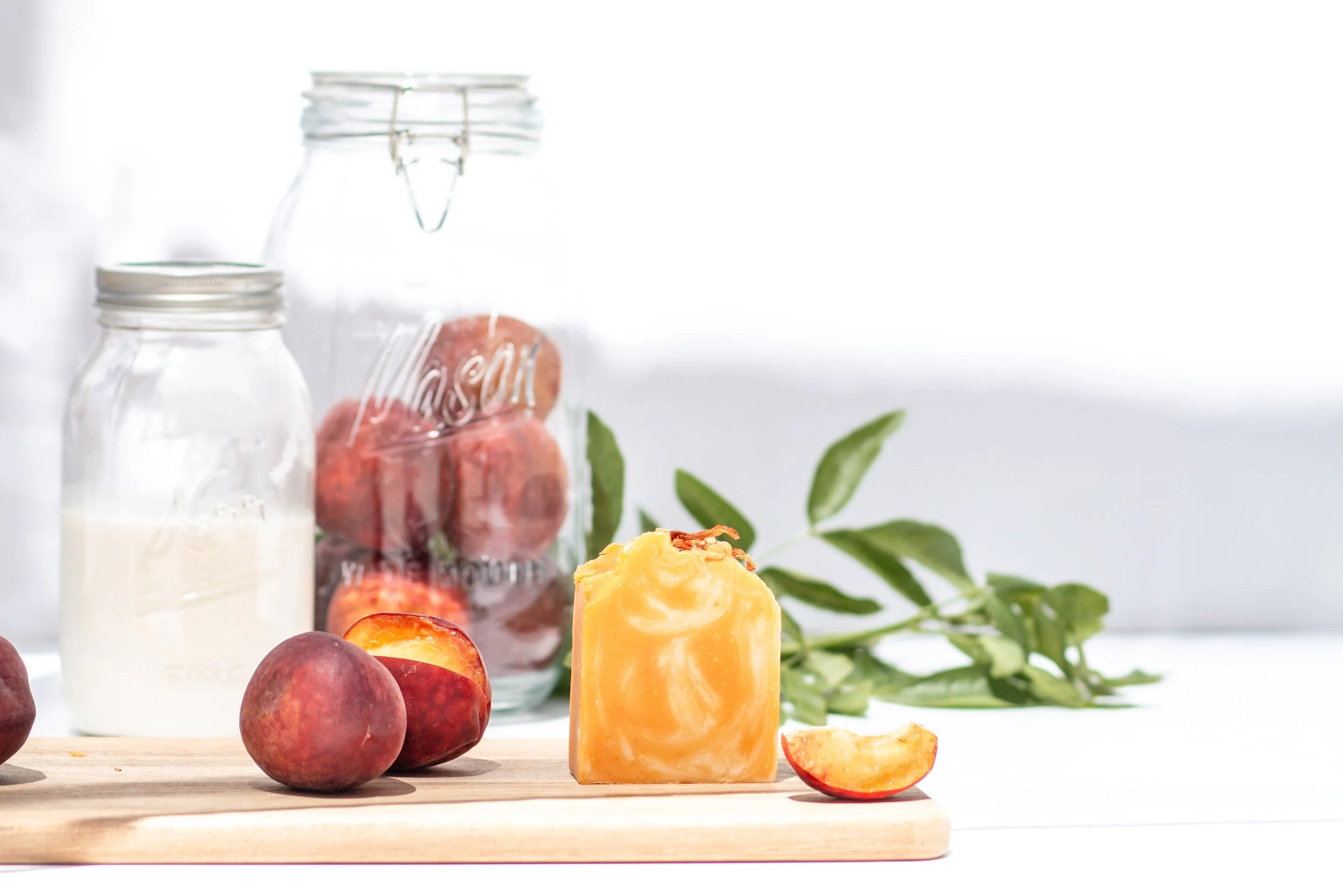 Fruits including peaches and peach slices, a bar of soap on a wooden board, a jar of milk, and a jar of peaches, with green leaves in the background