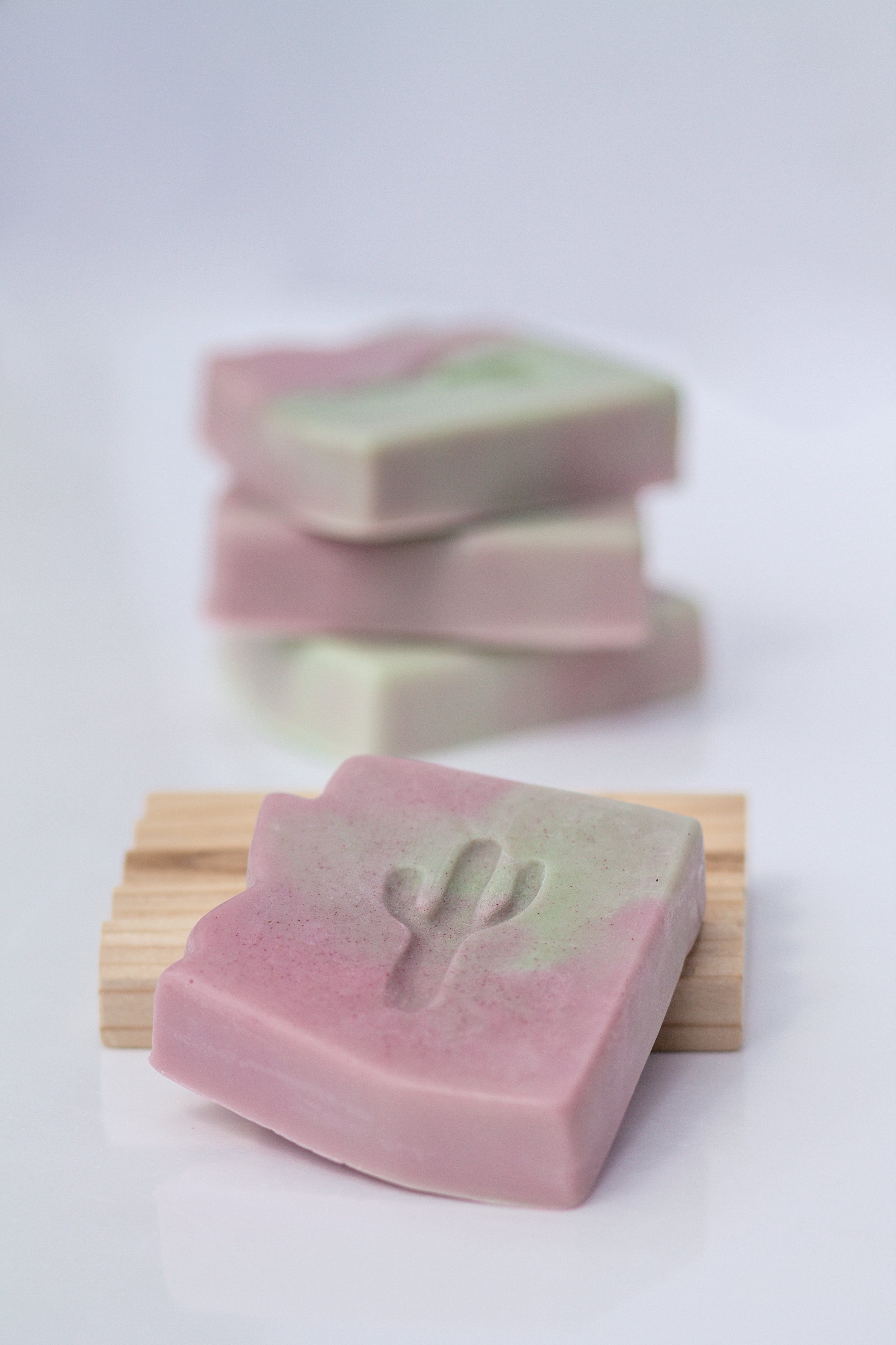 Pink and green soap with a cactus design embossed on it, placed on a small wooden platform with a stack of similar soaps in the background, all on a white surface.