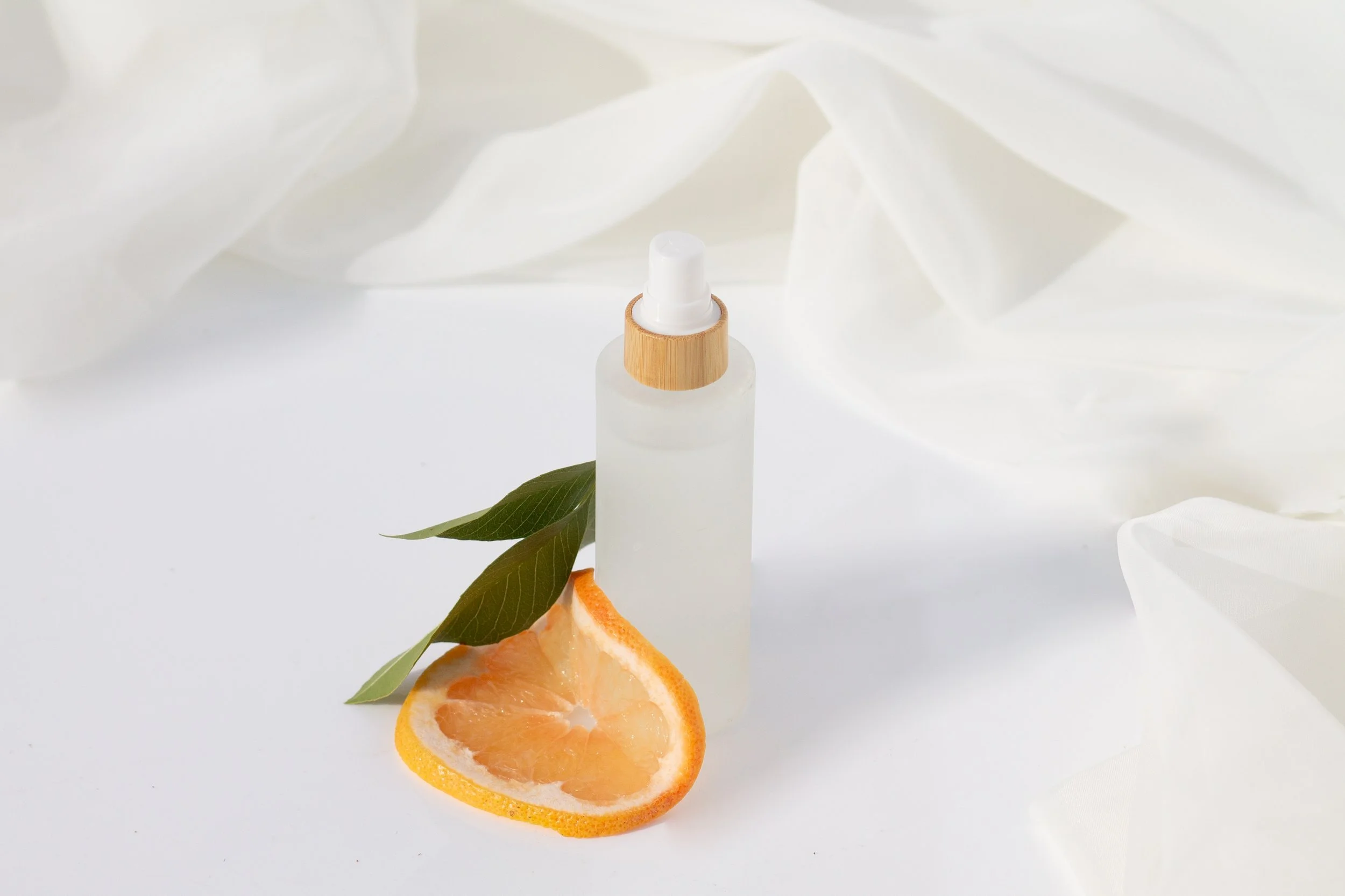 A frosted glass spray bottle with a white nozzle and a bamboo cap, next to a sliced orange and green leaves, on a white surface with soft white fabric in the background.