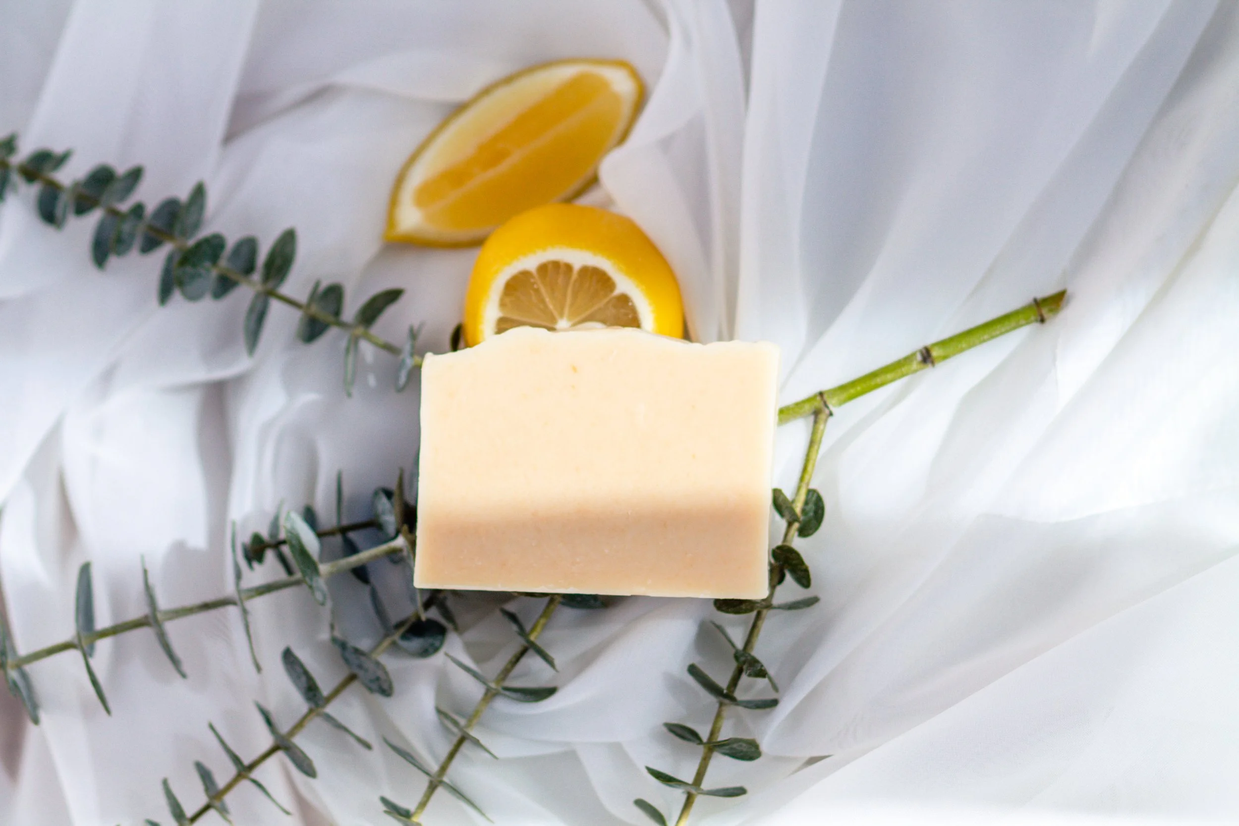 A bar of soap, lemon slices, and eucalyptus branches arranged on white fabric.