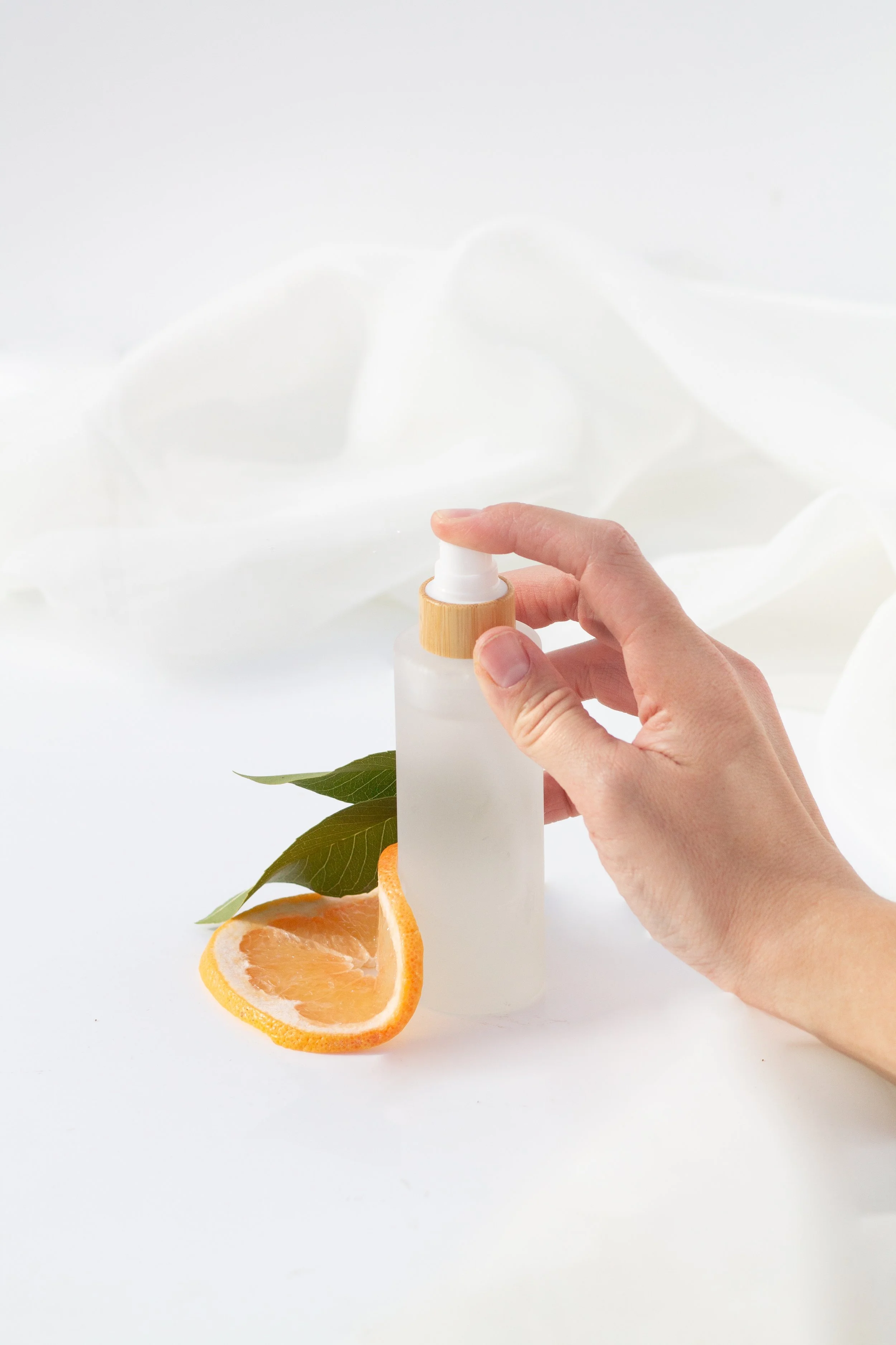 A person's hand holding a frosted glass spray bottle with a wooden cap, placed beside a sliced orange, green leaves, and a white textured fabric in the background.