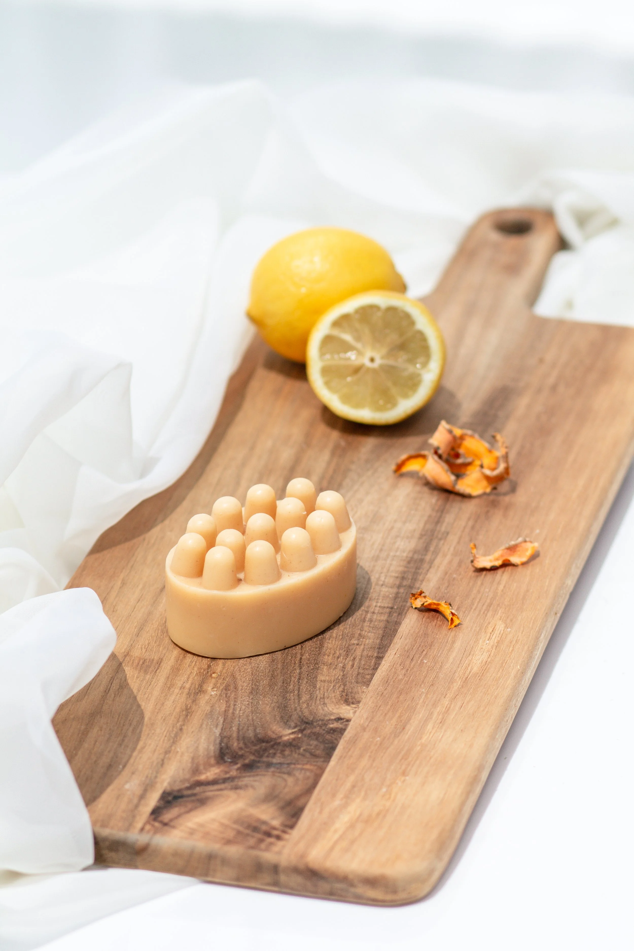 A wooden cutting board with lemon halves, a beige massage stone, and dried orange peel on it, against a white fabric background.