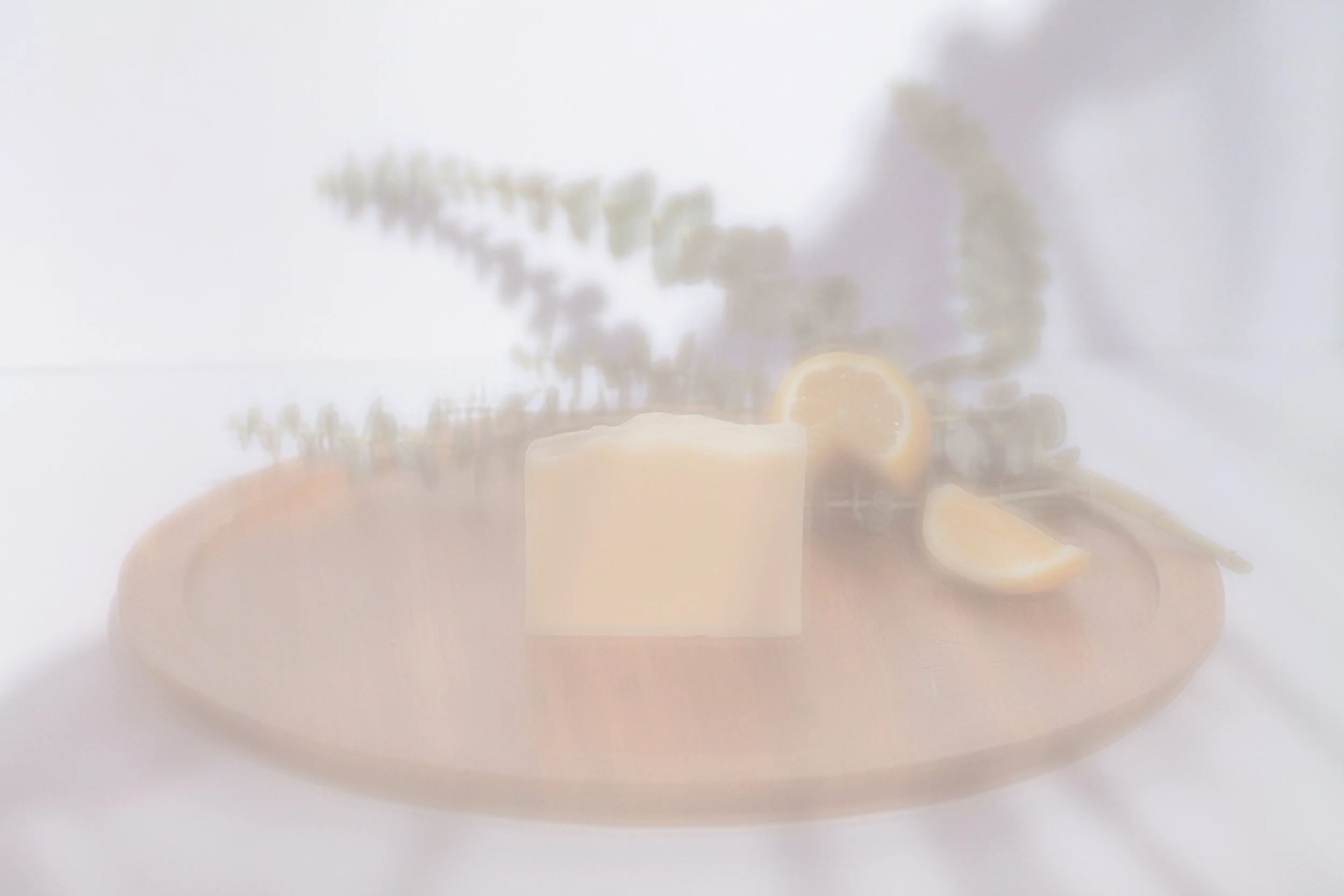 Blurred image of bar soap, lemon slices, and herbs on a wooden board.