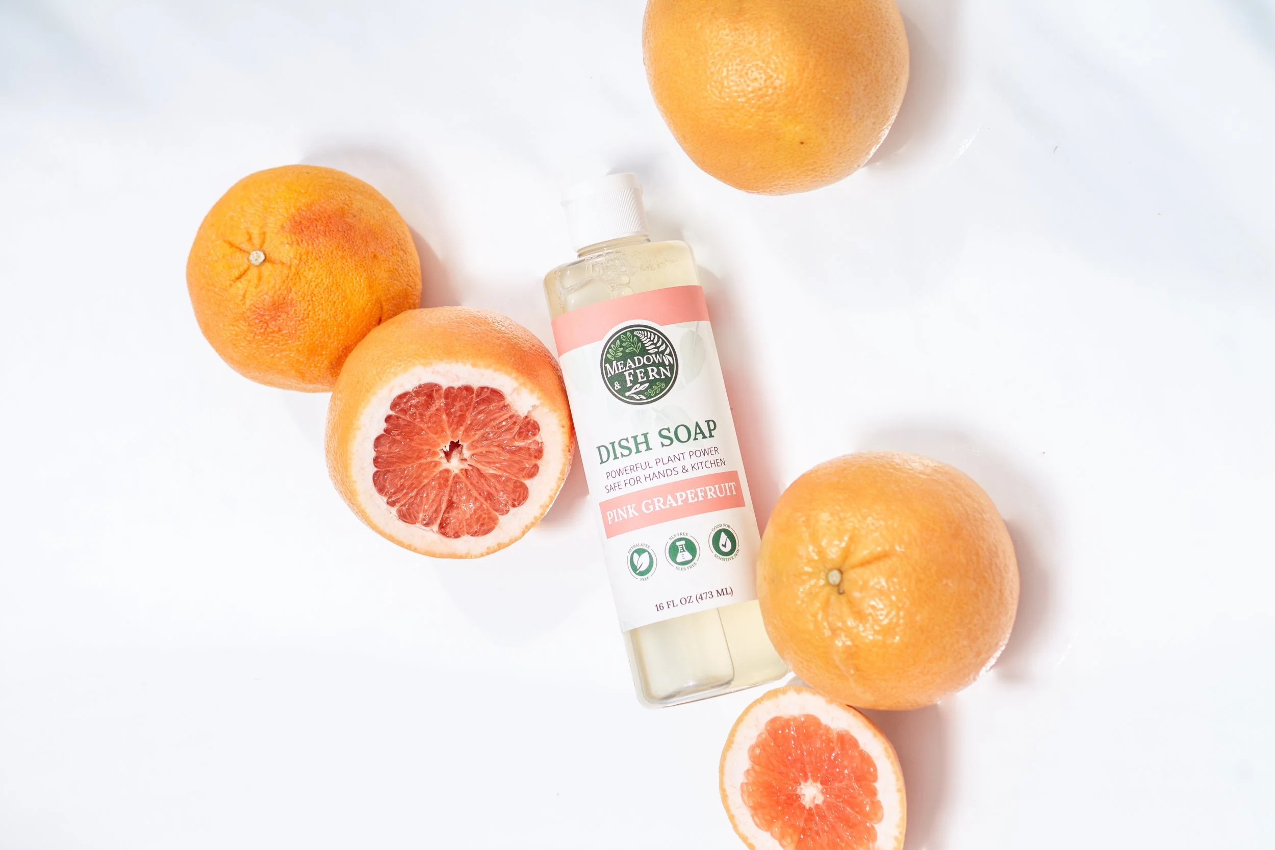 A bottle of Meadow & Fern dish soap with pink grapefruit scent surrounded by whole and halved grapefruits on a white surface.