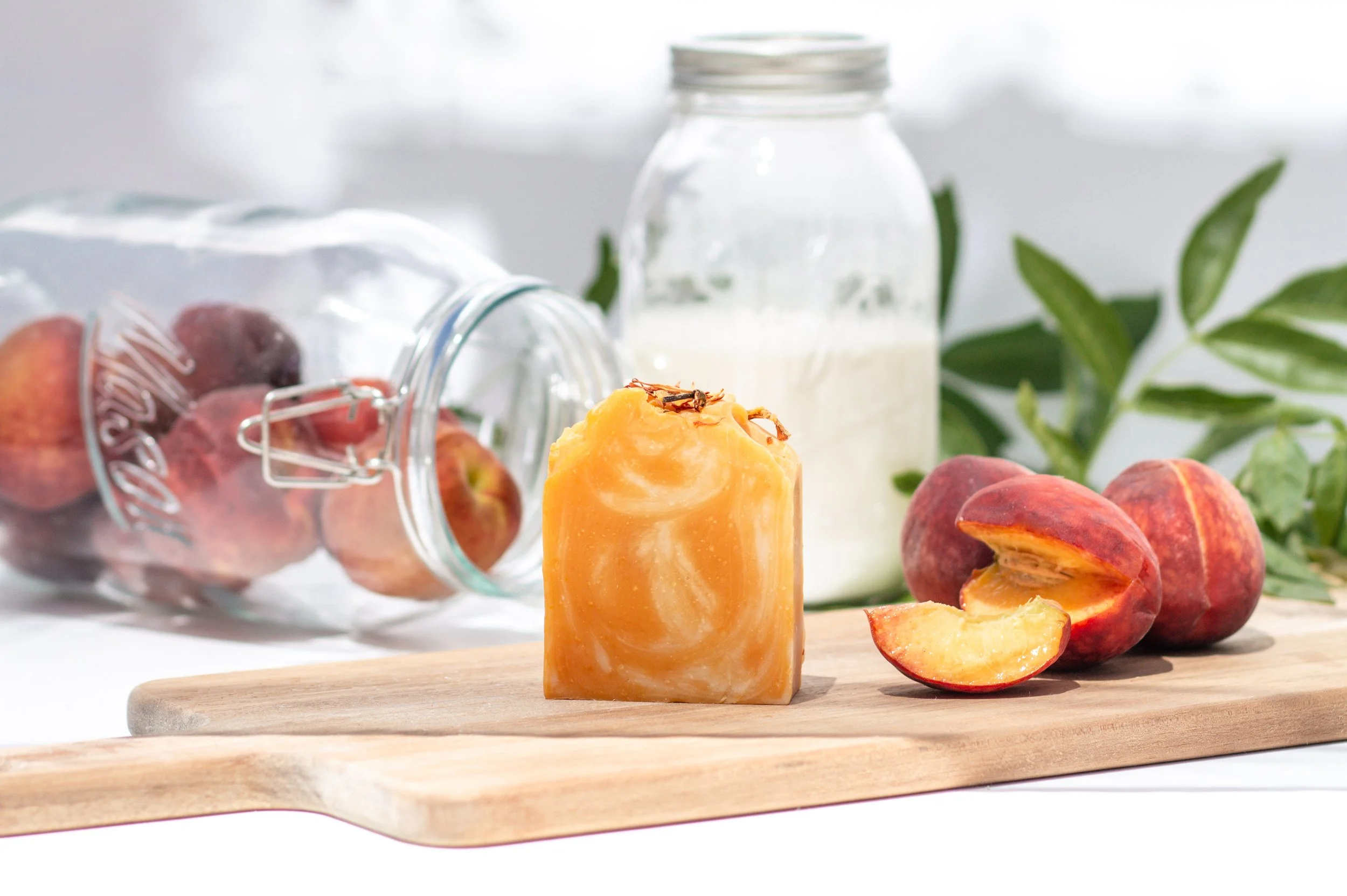 Bar of soap and peach slices on a wooden cutting board with a jar of milk, green leafy branch, and a glass jar of peaches in the background.