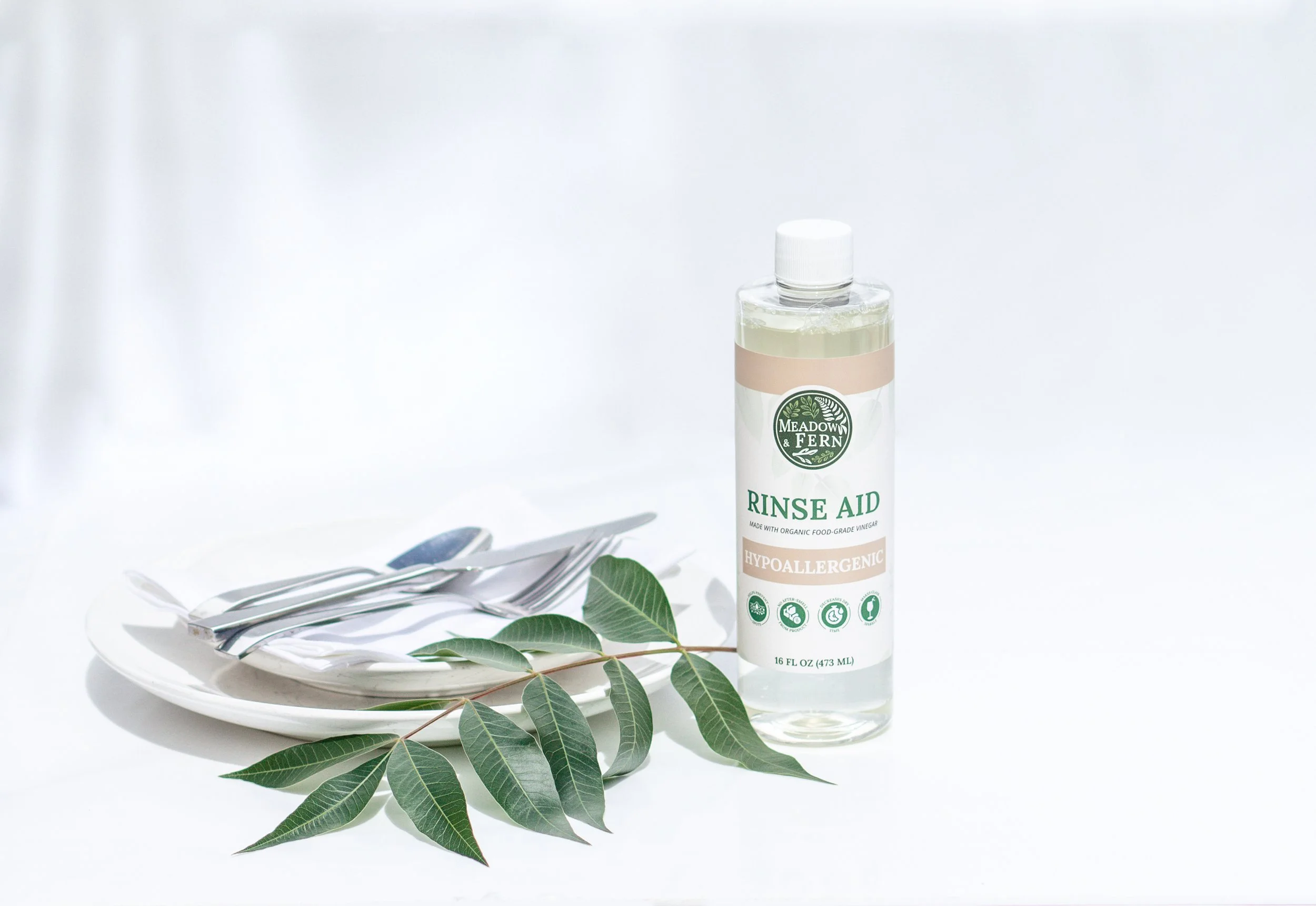 A bottle of Meadow & Fern rinse aid on a white surface next to a white plate with silverware and green leaves.