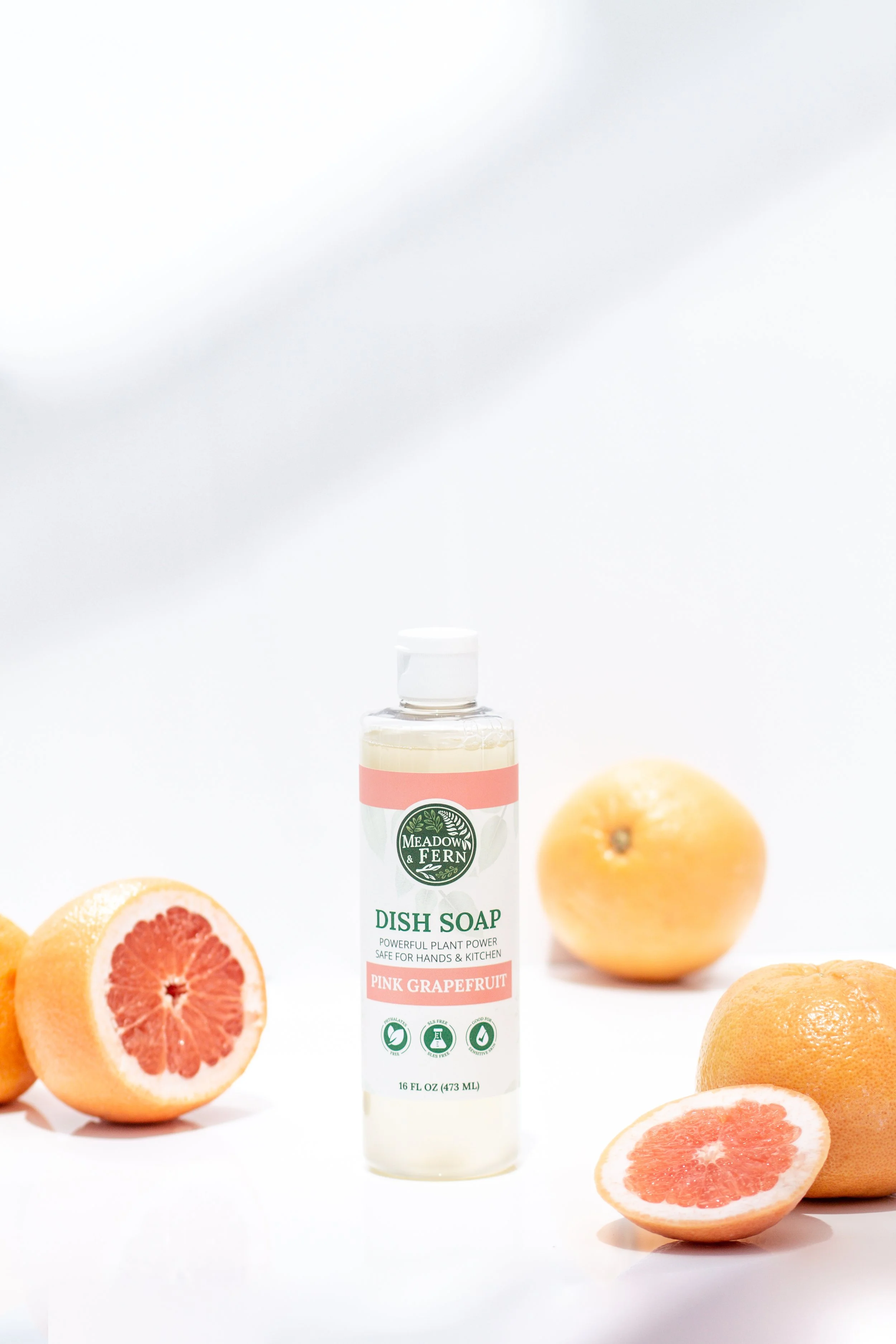 A bottle of Meadow & Fern pink grapefruit dish soap surrounded by whole and halved grapefruits on a white background.