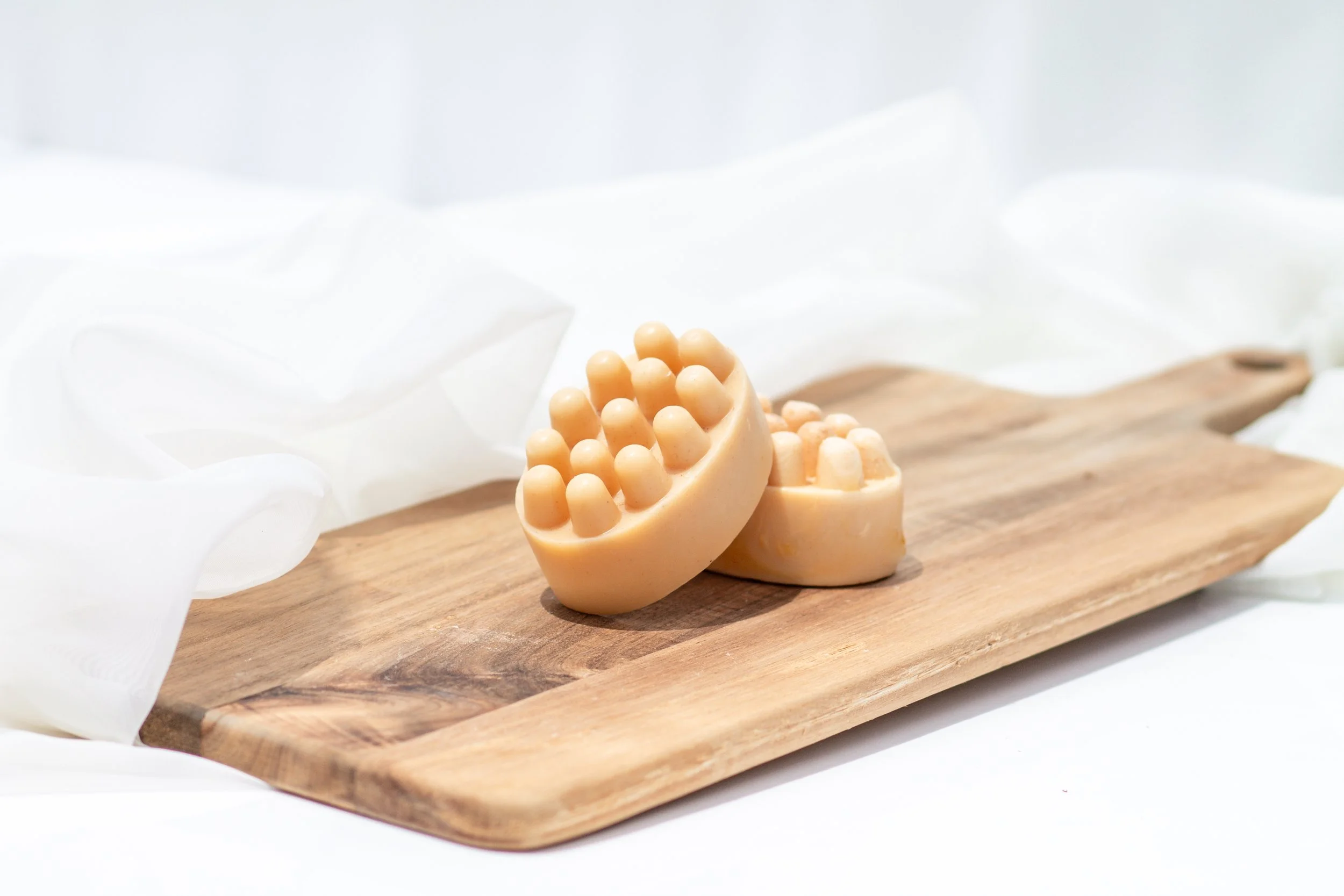 Two oval-shaped orange soap bars with textured tops on a wooden cutting board, surrounded by white fabric.