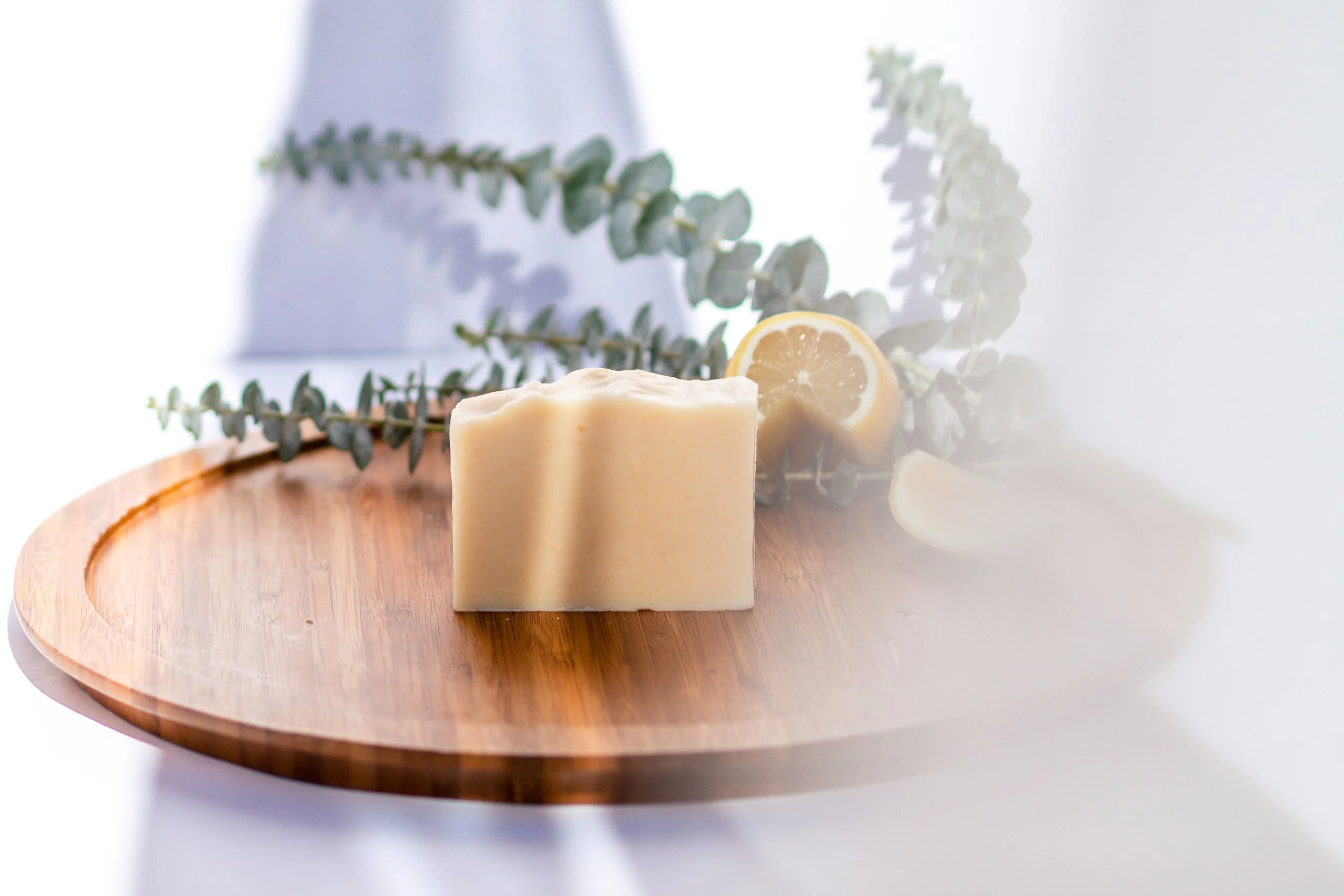 A bar of soap on a round wooden tray with lemon slices and eucalyptus leaves in the background.