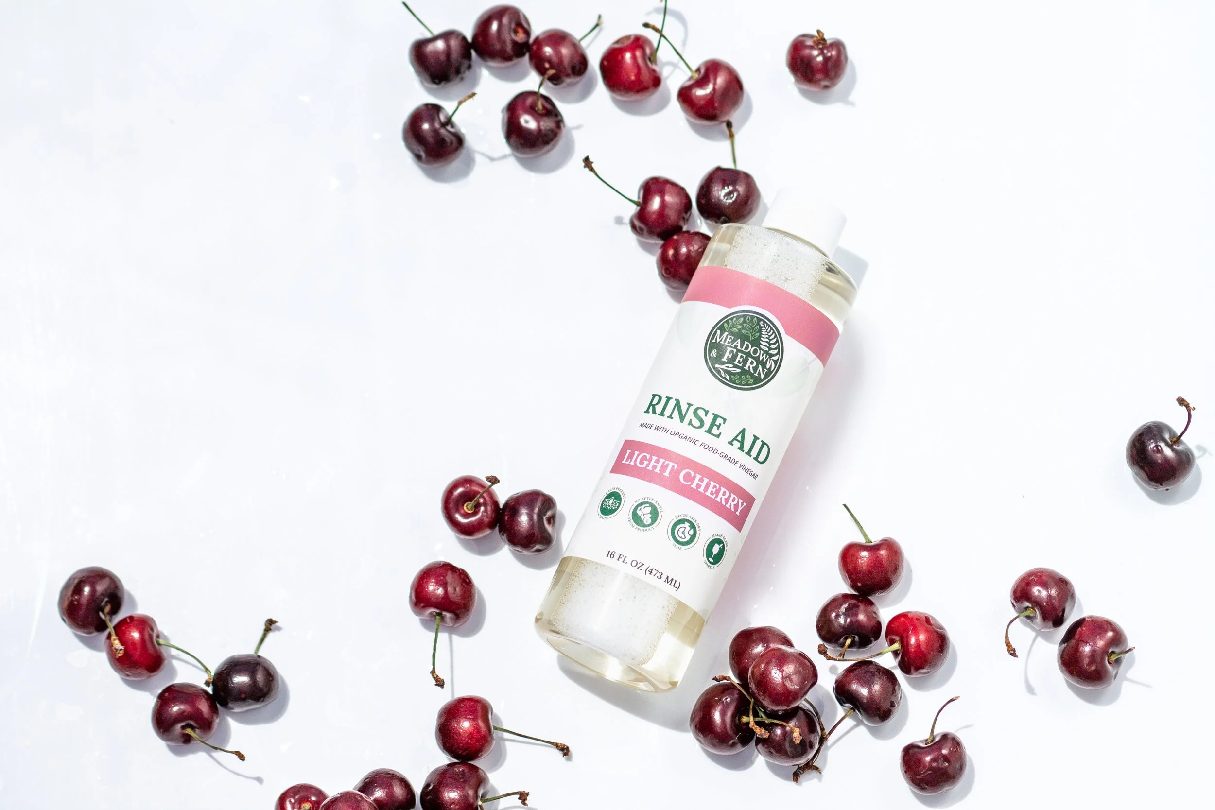 A bottle of Meadow & Fern rinse aid labeled 'Light Cherry' surrounded by scattered fresh dark red cherries on a white background.