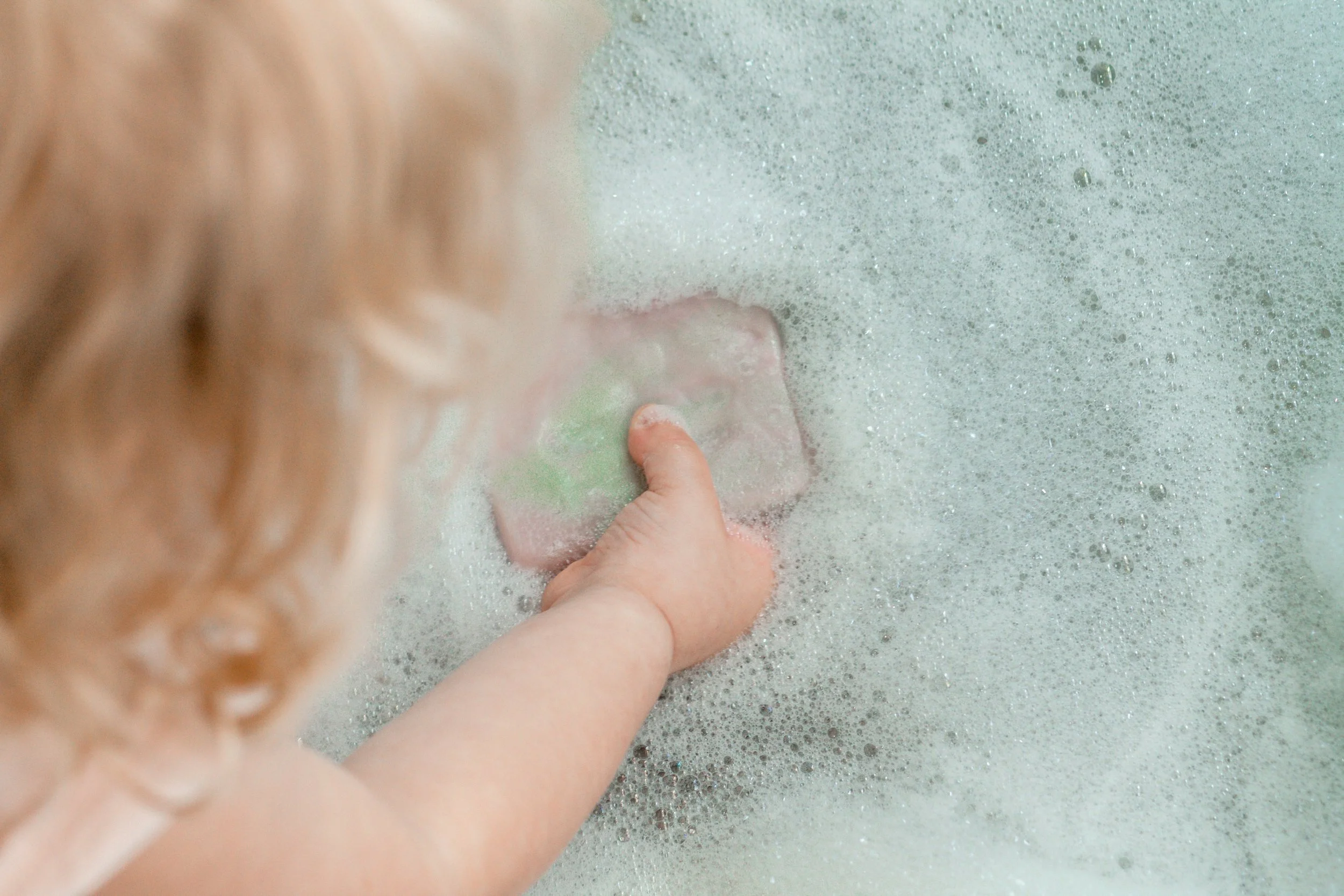 Child playing with a pink and green bar of soap in a bubbly kitchen sink.