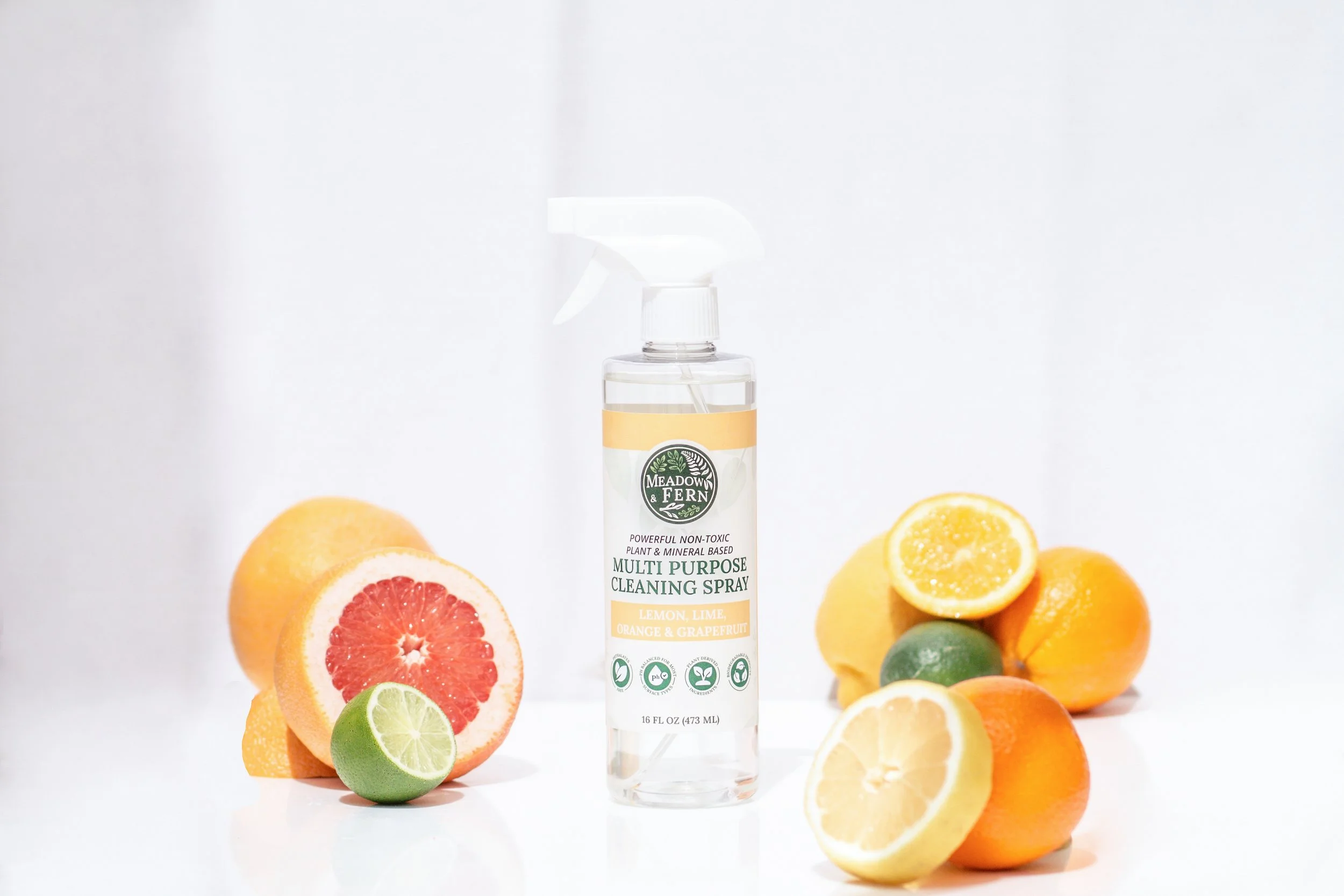 A spray bottle of Meadow & Fern multi purpose cleaning spray surrounded by sliced citrus fruits including lemon, lime, orange, grapefruit, and lemon