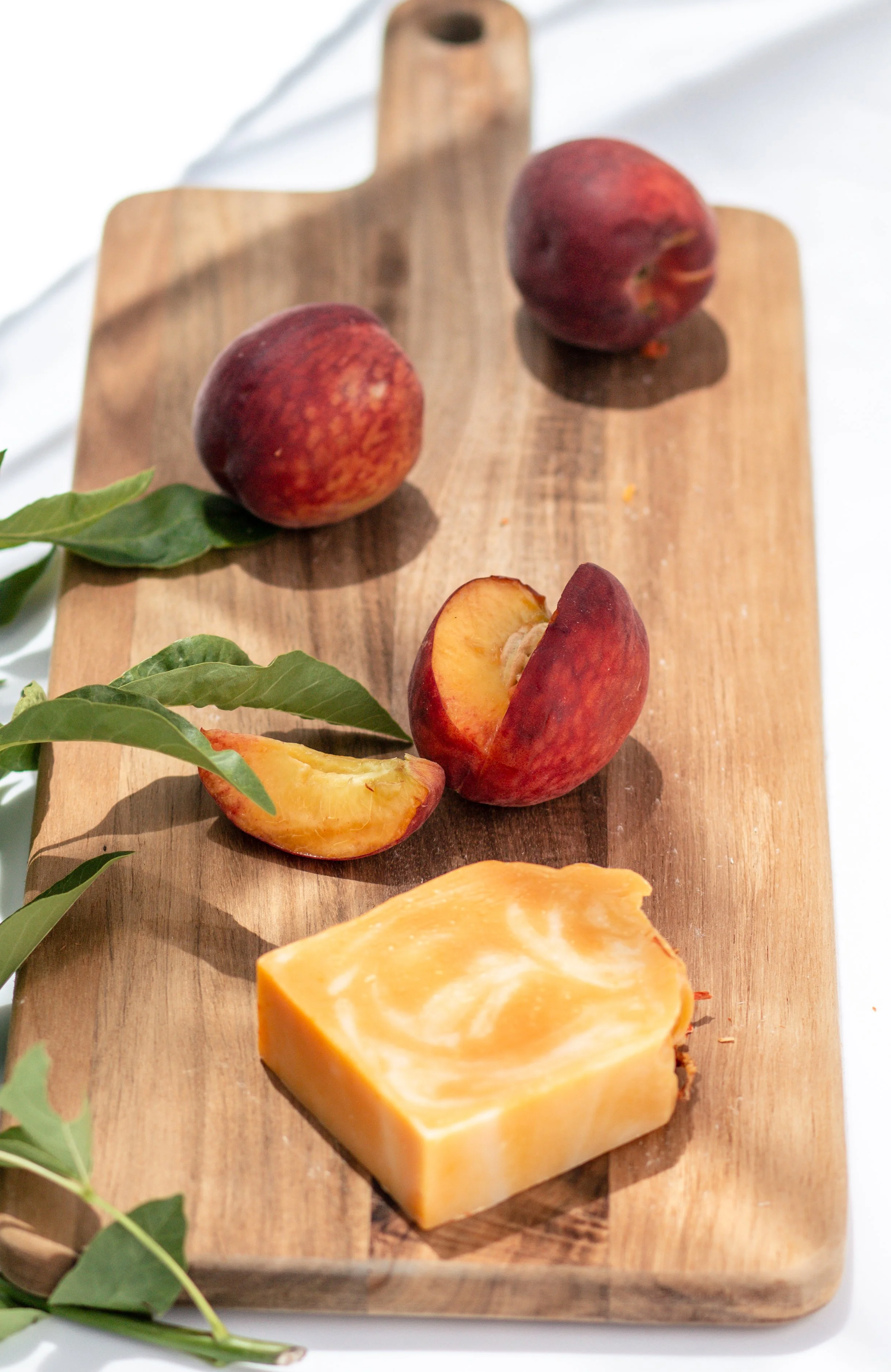 Three whole peaches, one sliced peach, and a bar of soap on a wooden cutting board, with green leaves nearby.