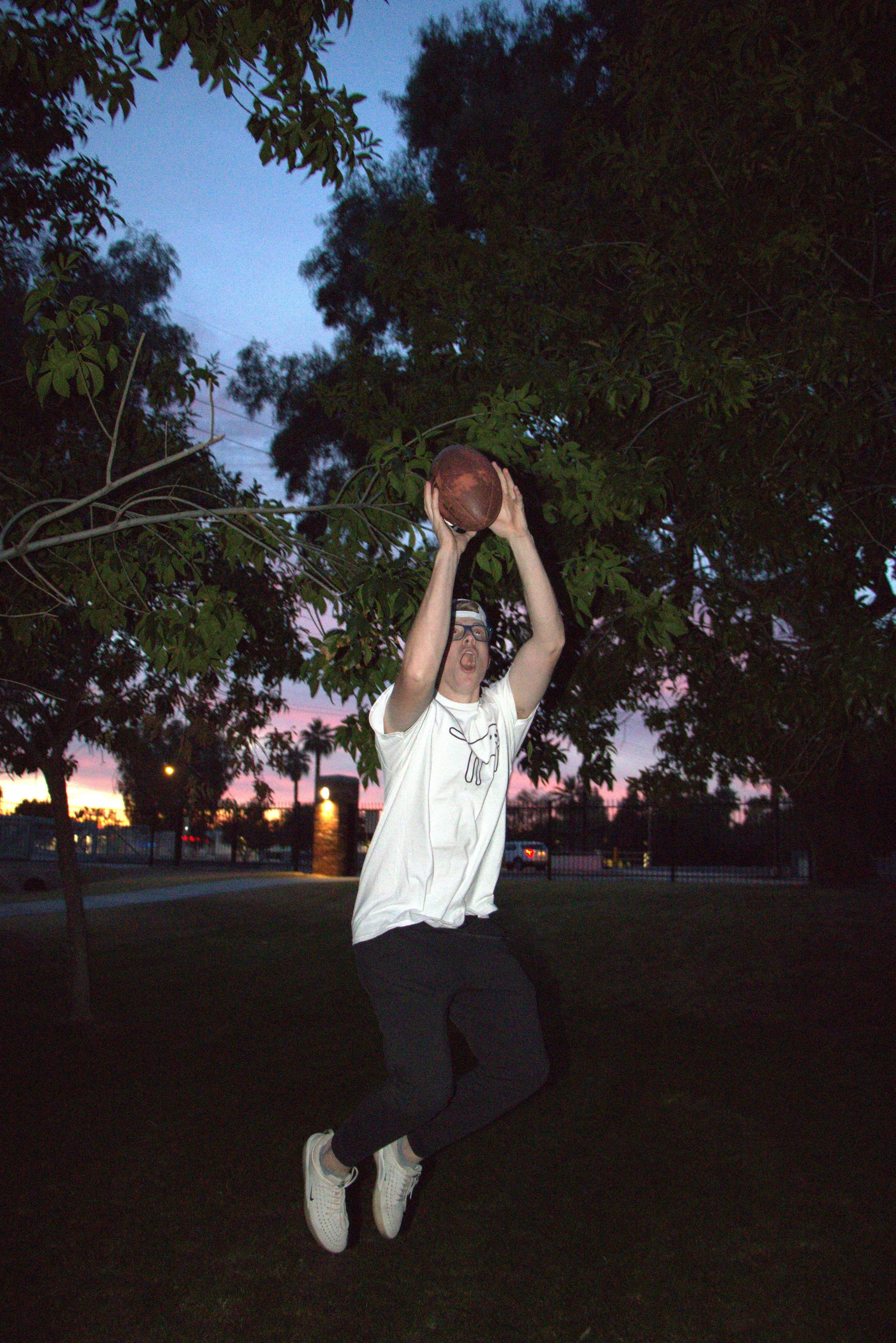 A person jumping in the air while holding a basketball, playing basketball outdoors during sunset.