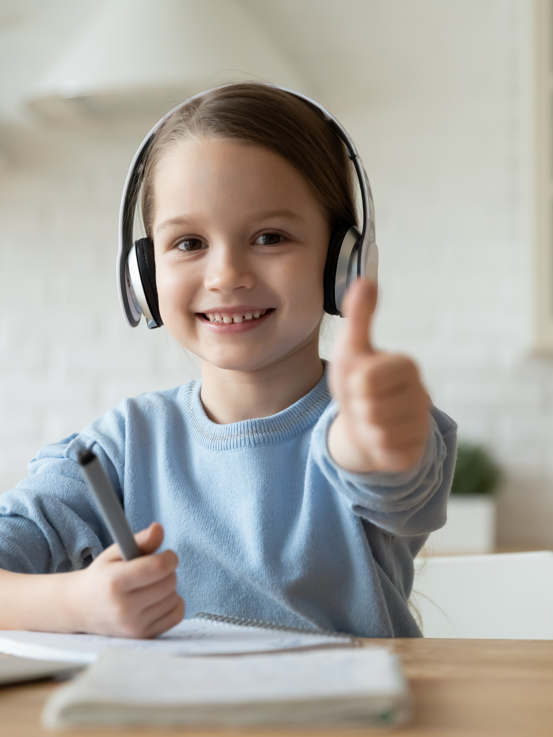 A smiling young girl wearing headphones giving a thumbs-up while sitting at a table with a notebook and pen.