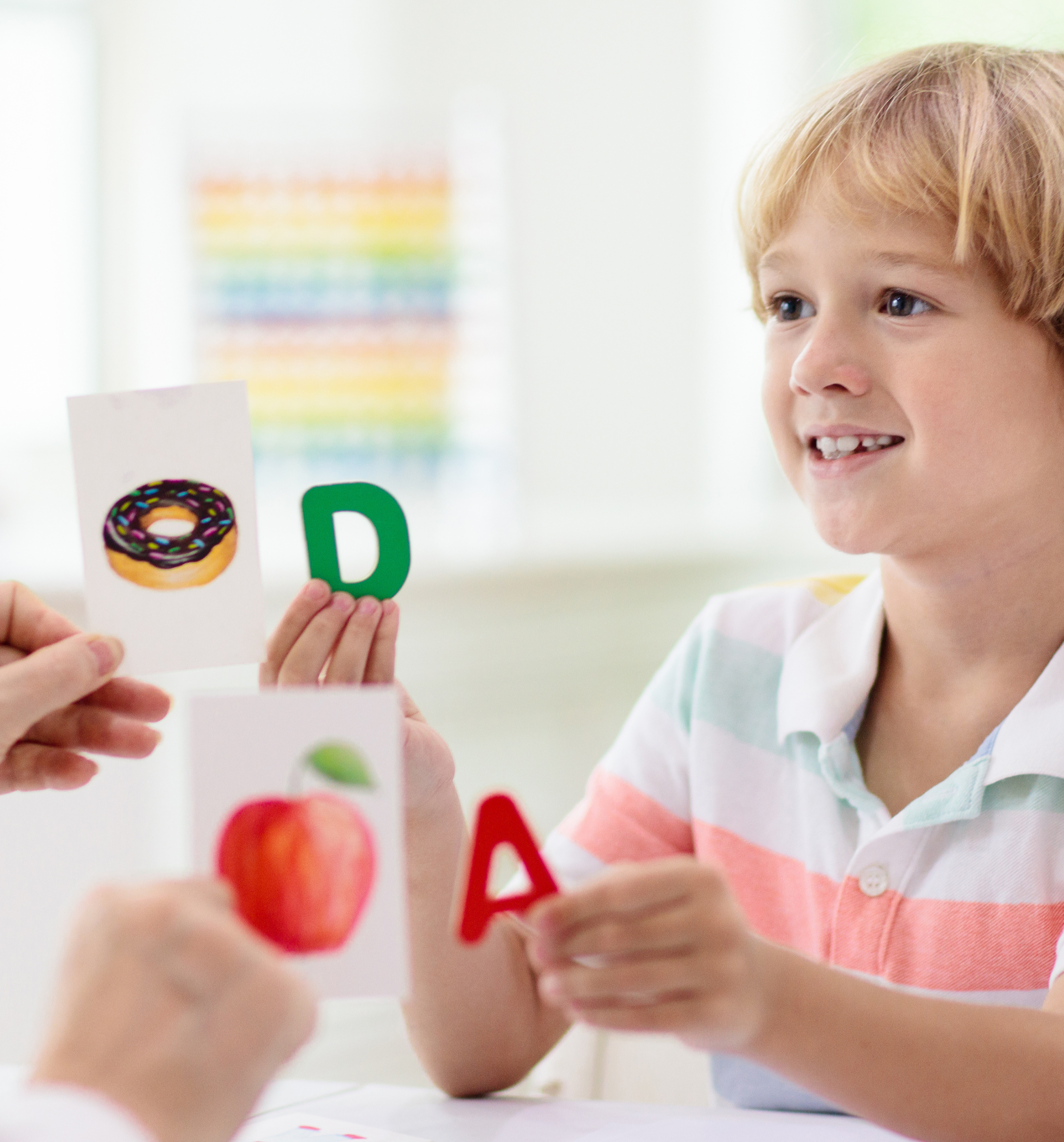 A young boy with blonde hair participating in an educational activity with flashcards, including images of a donut and an apple, and letters D and A.