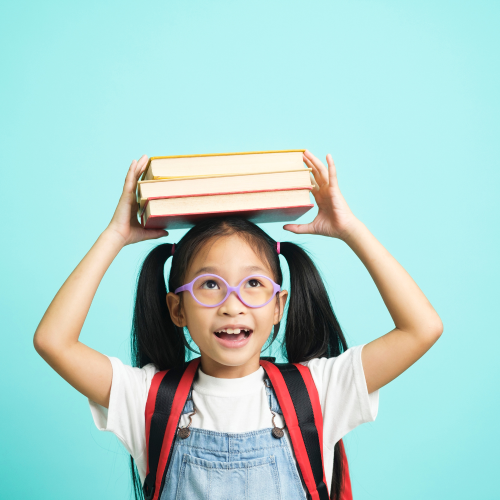 A young girl with glasses and pigtails, carrying a backpack, balancing four books on her head, smiling against a light blue background.