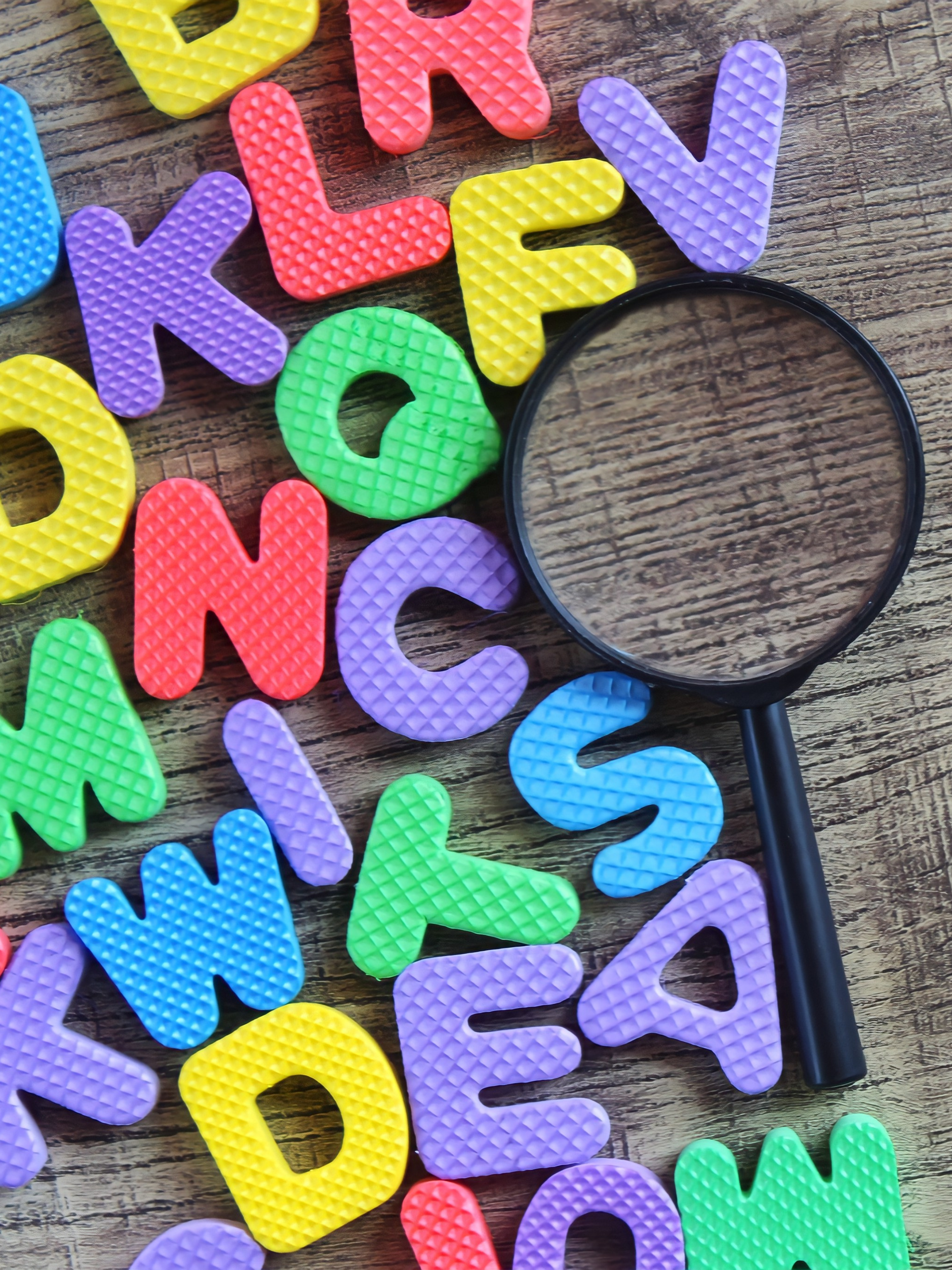 Colorful plastic alphabet letters scattered on a wooden surface with a magnifying glass beside them.
