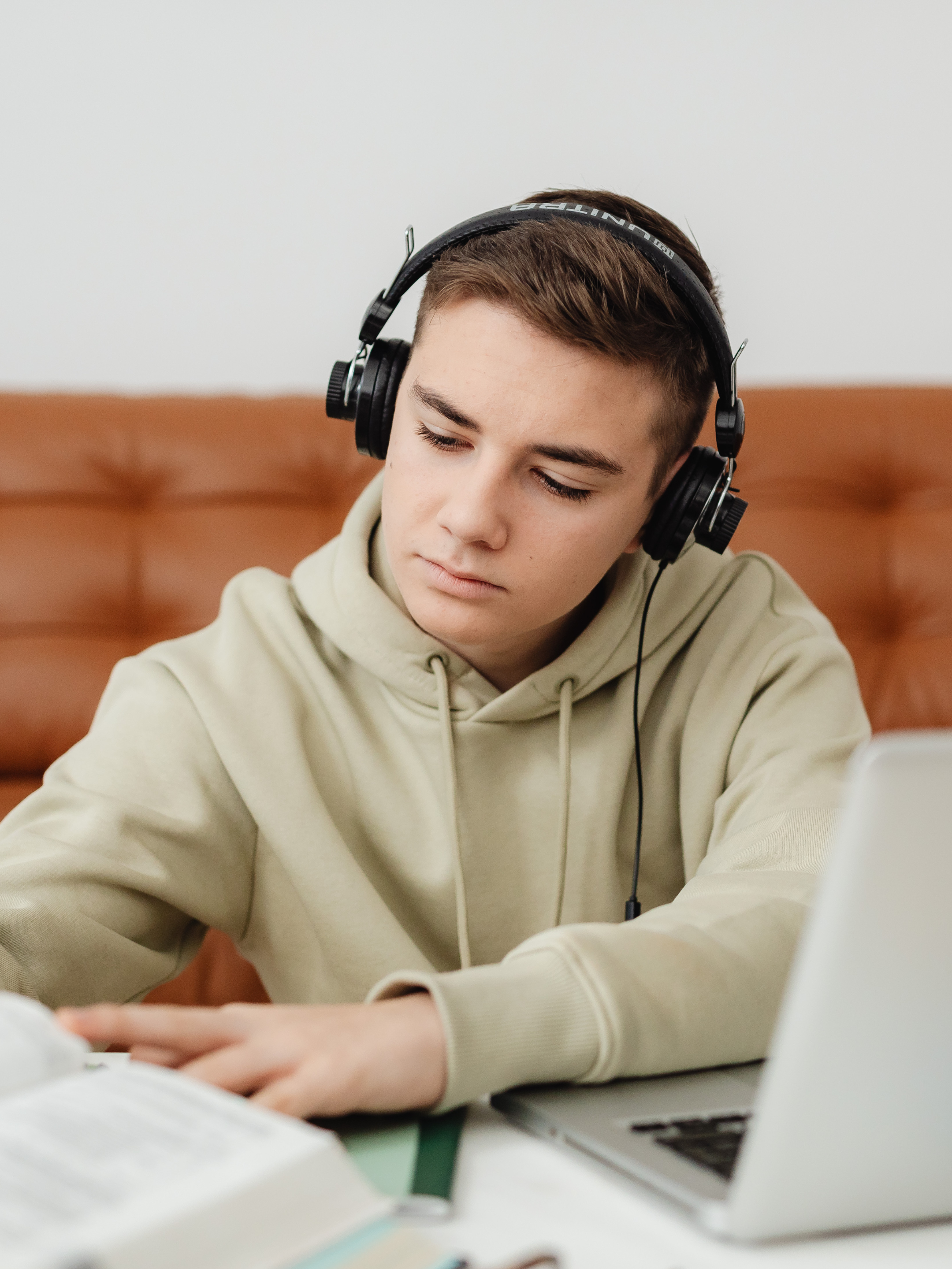 Young adult with short brown hair wearing headphones, learning studying or working at a desk with books, a notebook, and a laptop.